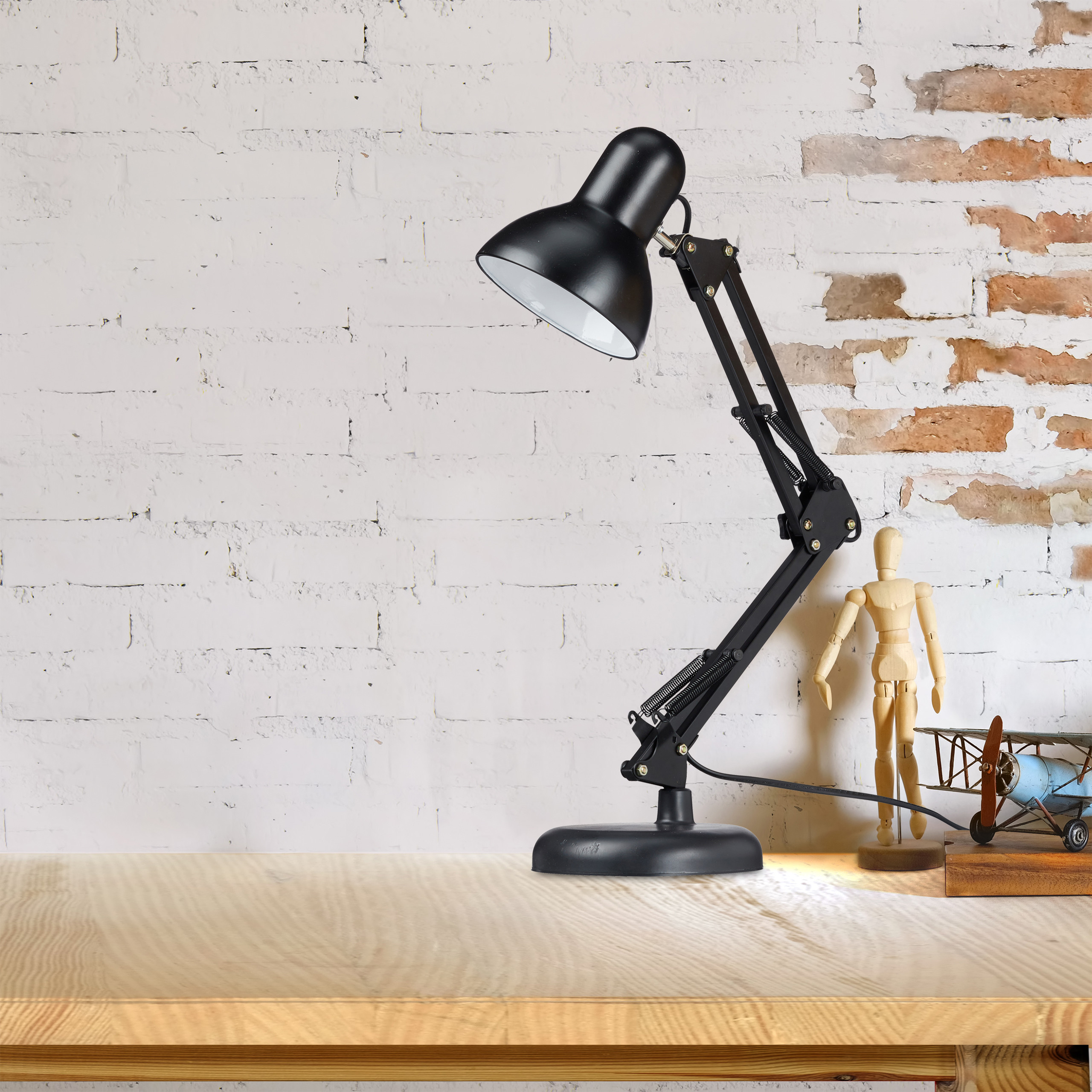 Eine schwarze Schreibtischlampe, eine Holzpuppe und ein Spielzeugflugzeug stehen auf einem Holztisch vor einer Backsteinwand.