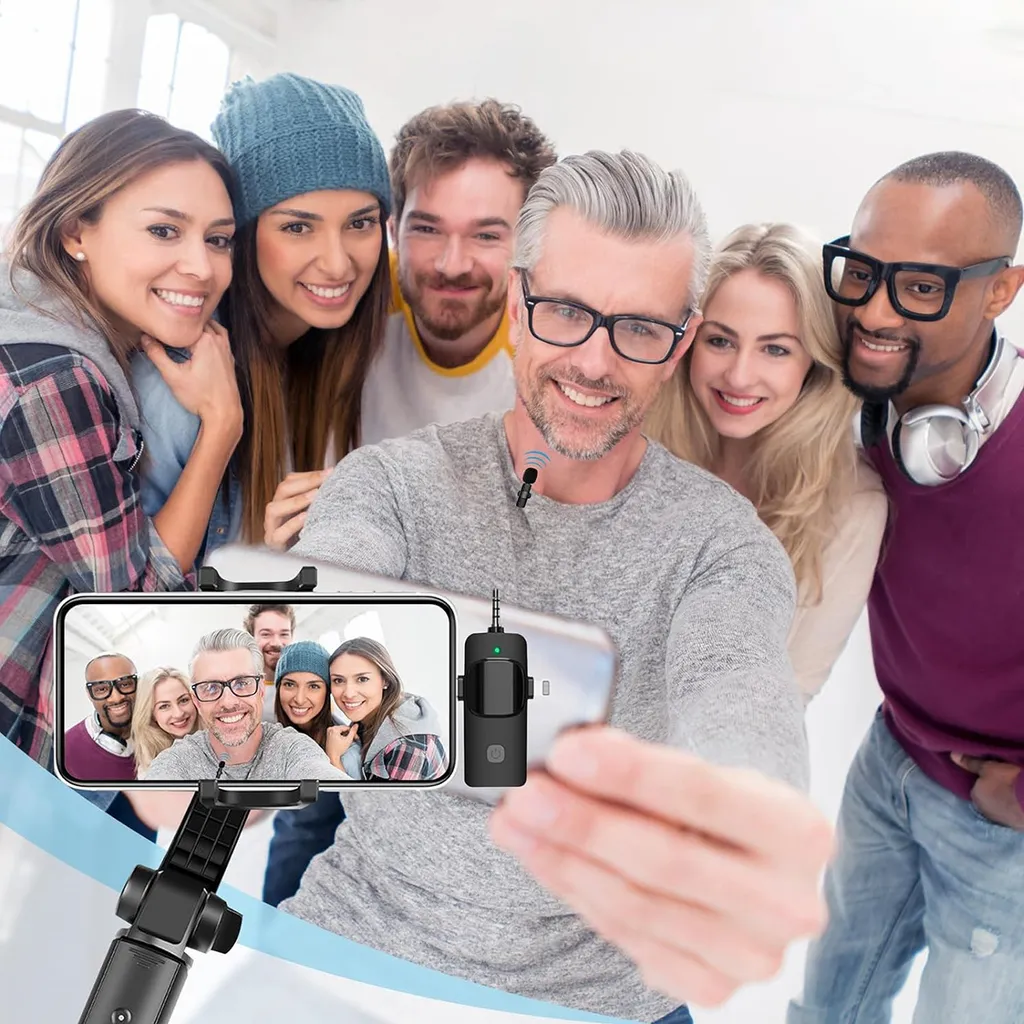 Sechs diverse Freunde lächeln, während ein Mann im Vordergrund ein Selfie mit einem Smartphone an einem Stick macht.