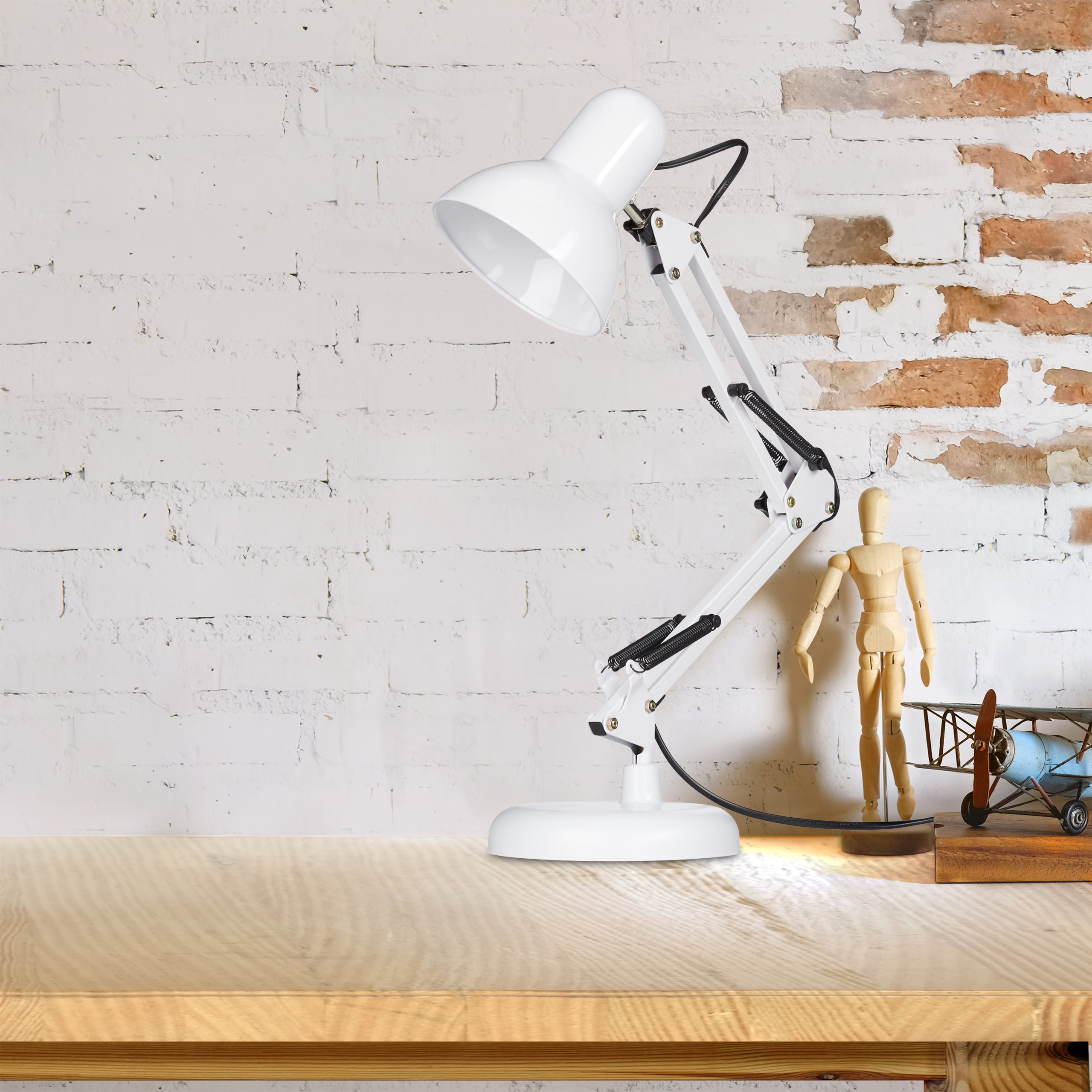 Eine weiße Schreibtischlampe, eine Holzpuppe und ein kleines Flugzeug stehen auf einem Holztisch vor einer Backsteinwand.