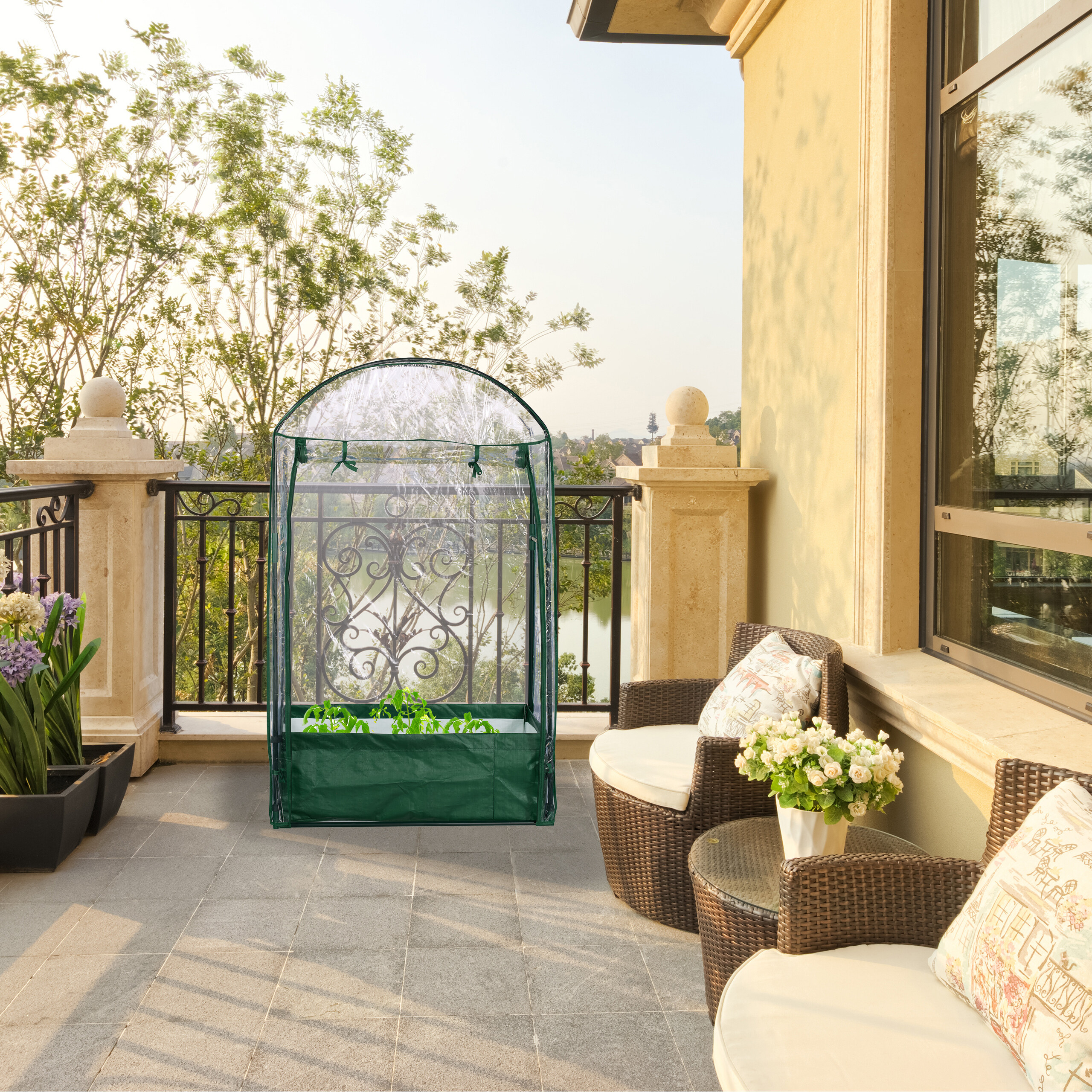 Ein kleines grünes Gewächshaus steht auf einer sonnigen Terrasse neben Gartenmöbeln mit Blick auf Bäume.