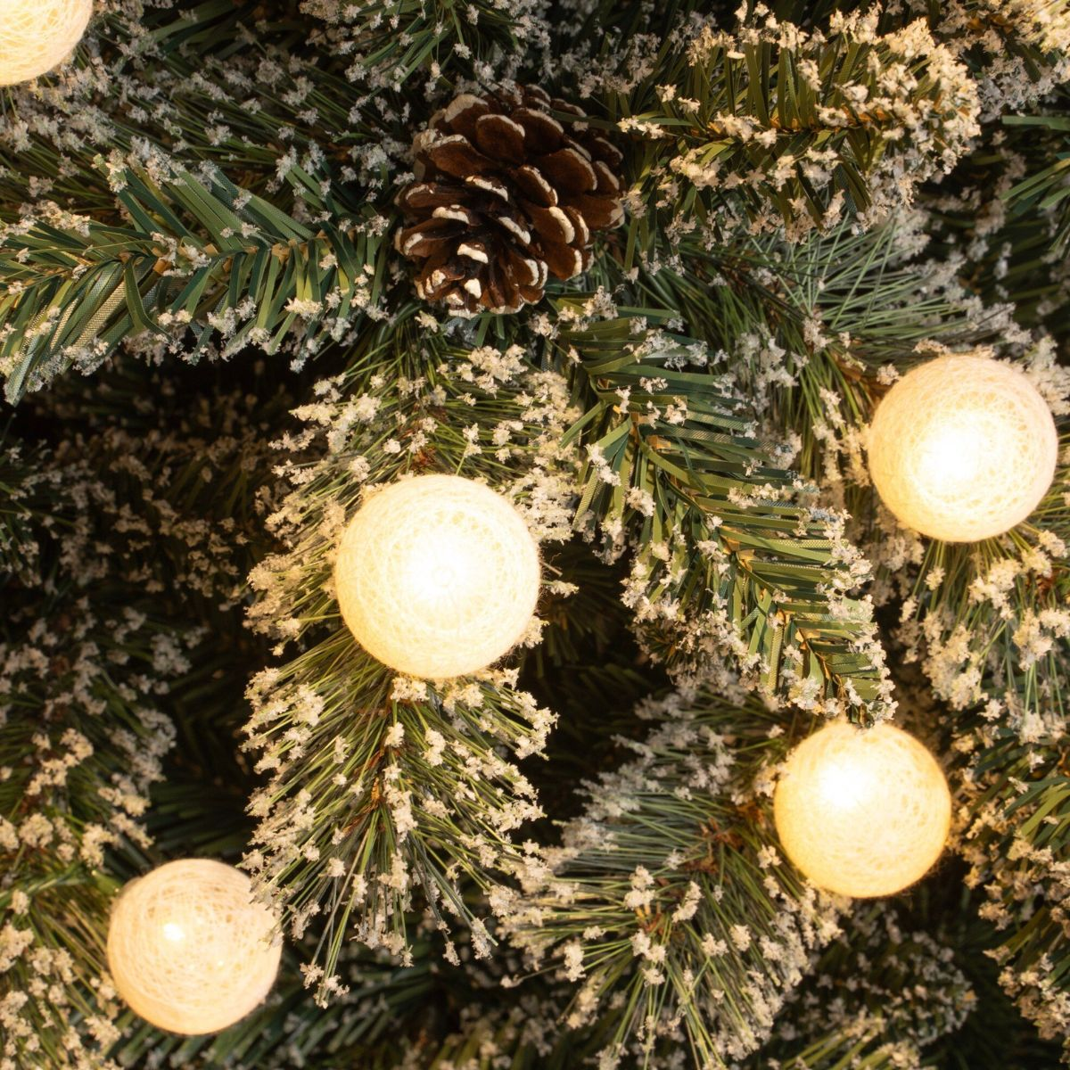 Besneeuwde kerstboom met gloeiende lampjes en dennenappel.