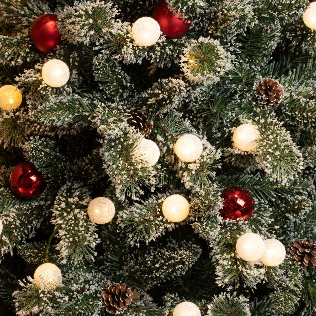 Besneeuwde kerstboom versierd met rode kerstballen, dennenappels en warme lampjes.