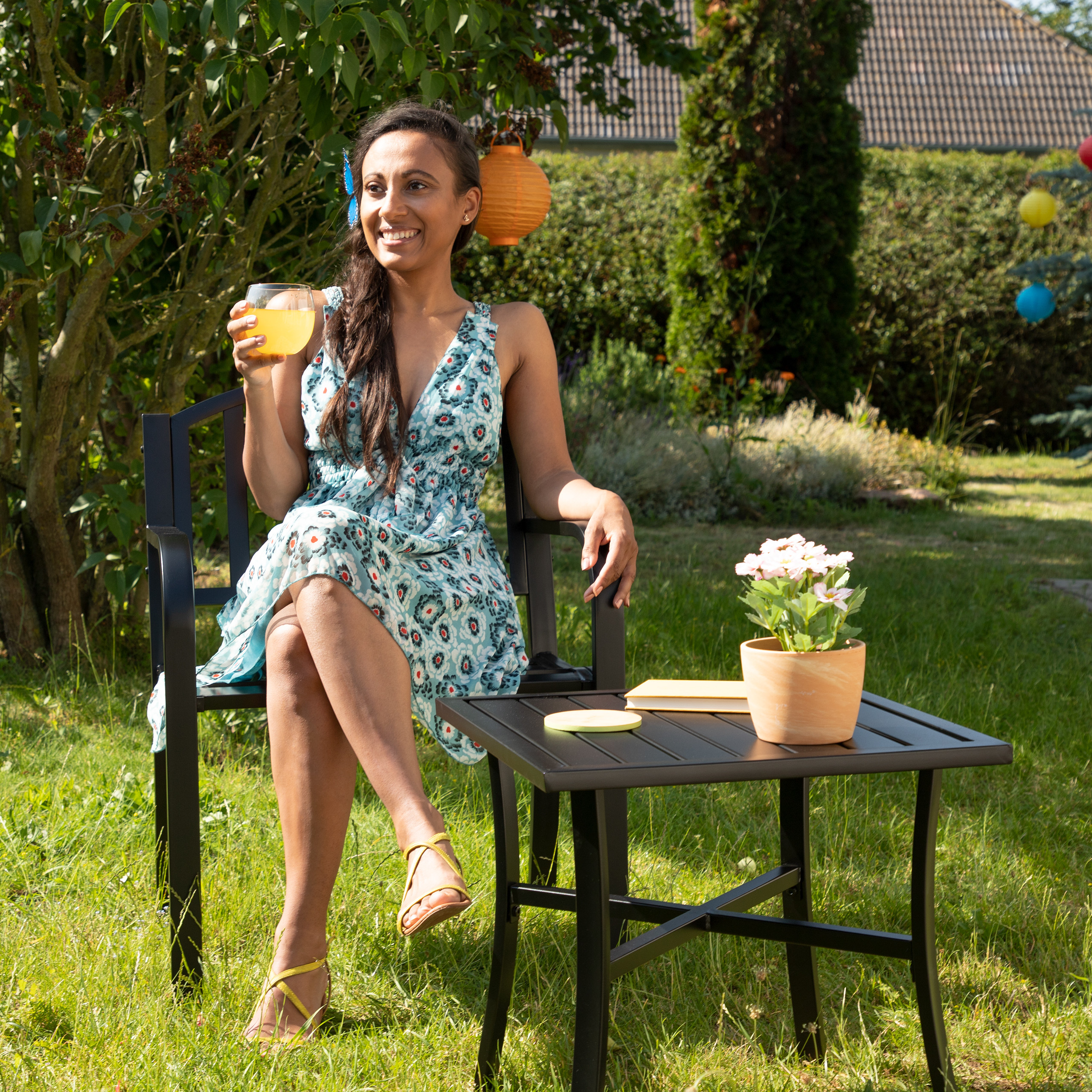 Eine Frau in einem blau-gemusterten Sommerkleid sitzt im Garten und hält ein Glas Orangensaft.