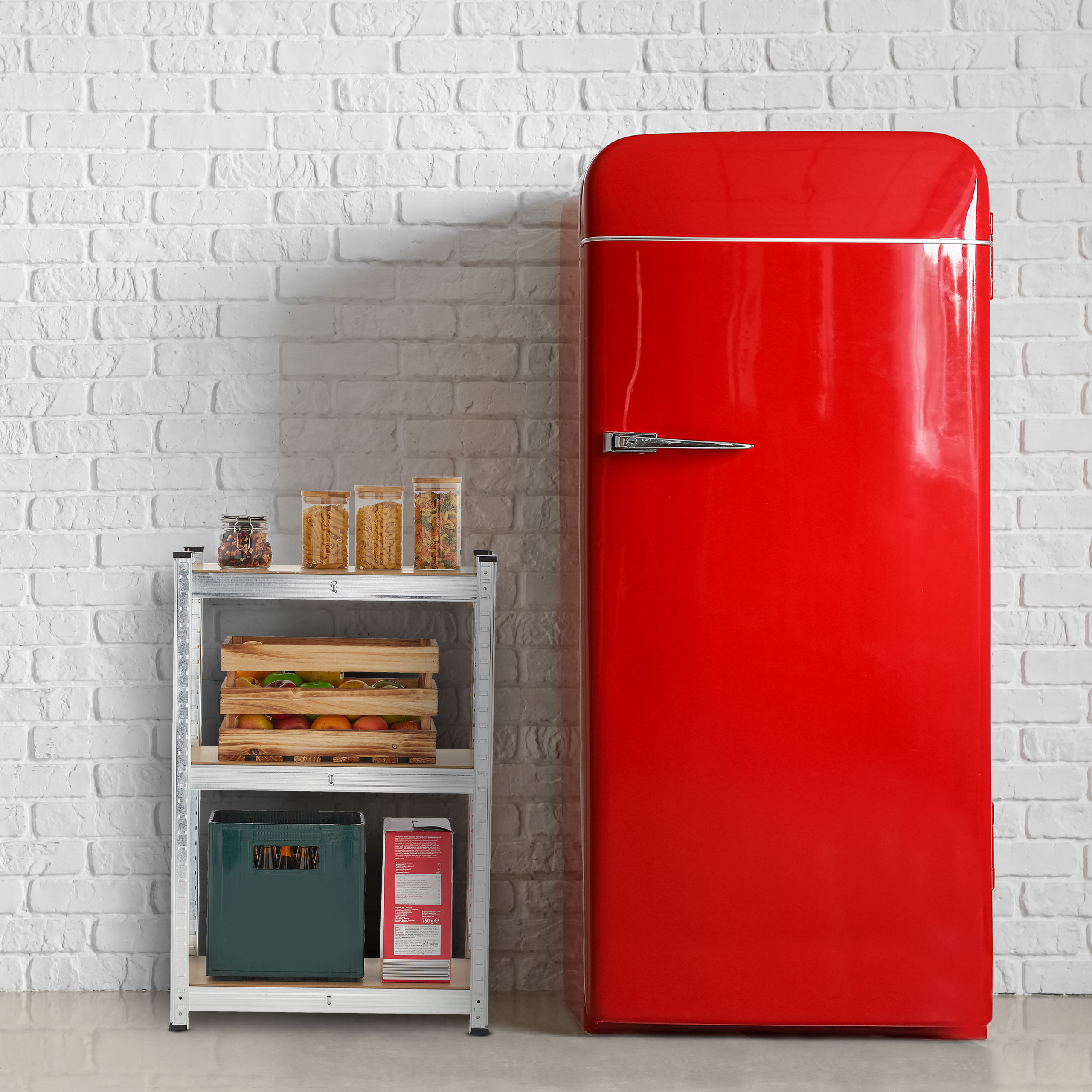Ein roter Retro-Kühlschrank steht neben einem Metallregal mit Vorratsgläsern und Kisten vor einer weißen Backsteinwand.