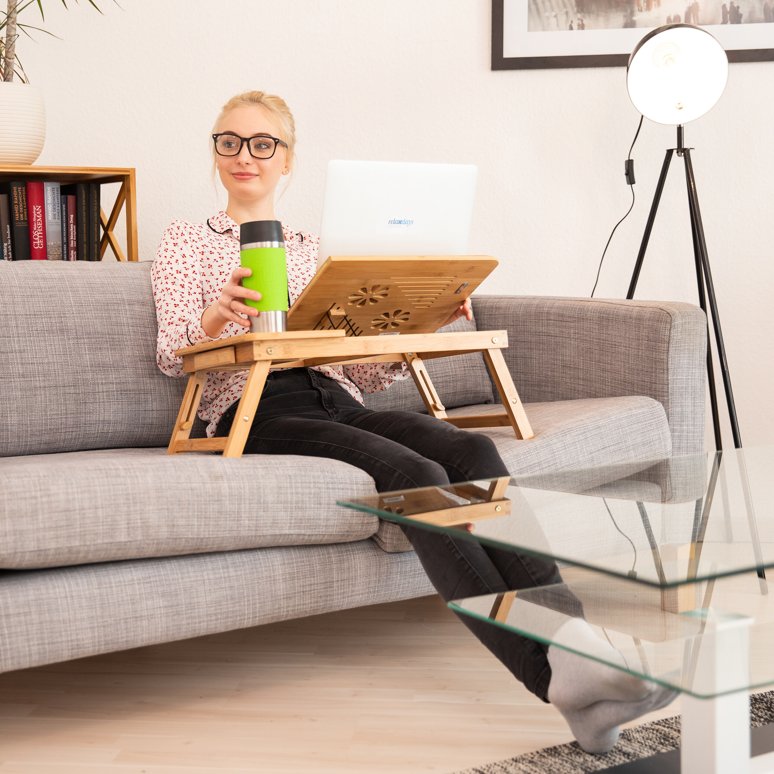 Eine blonde Frau sitzt auf einem grauen Sofa, arbeitet am Laptop und hält einen grünen Becher.