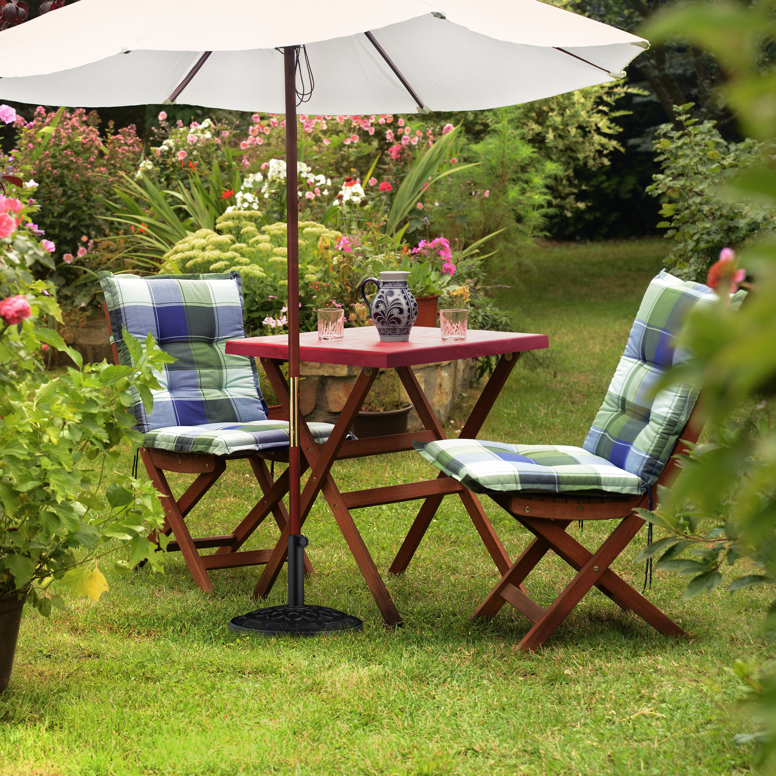 Ein Holztisch und zwei Stühle mit blau-grünen Kissen in einem Garten unter einem weißen Sonnenschirm.