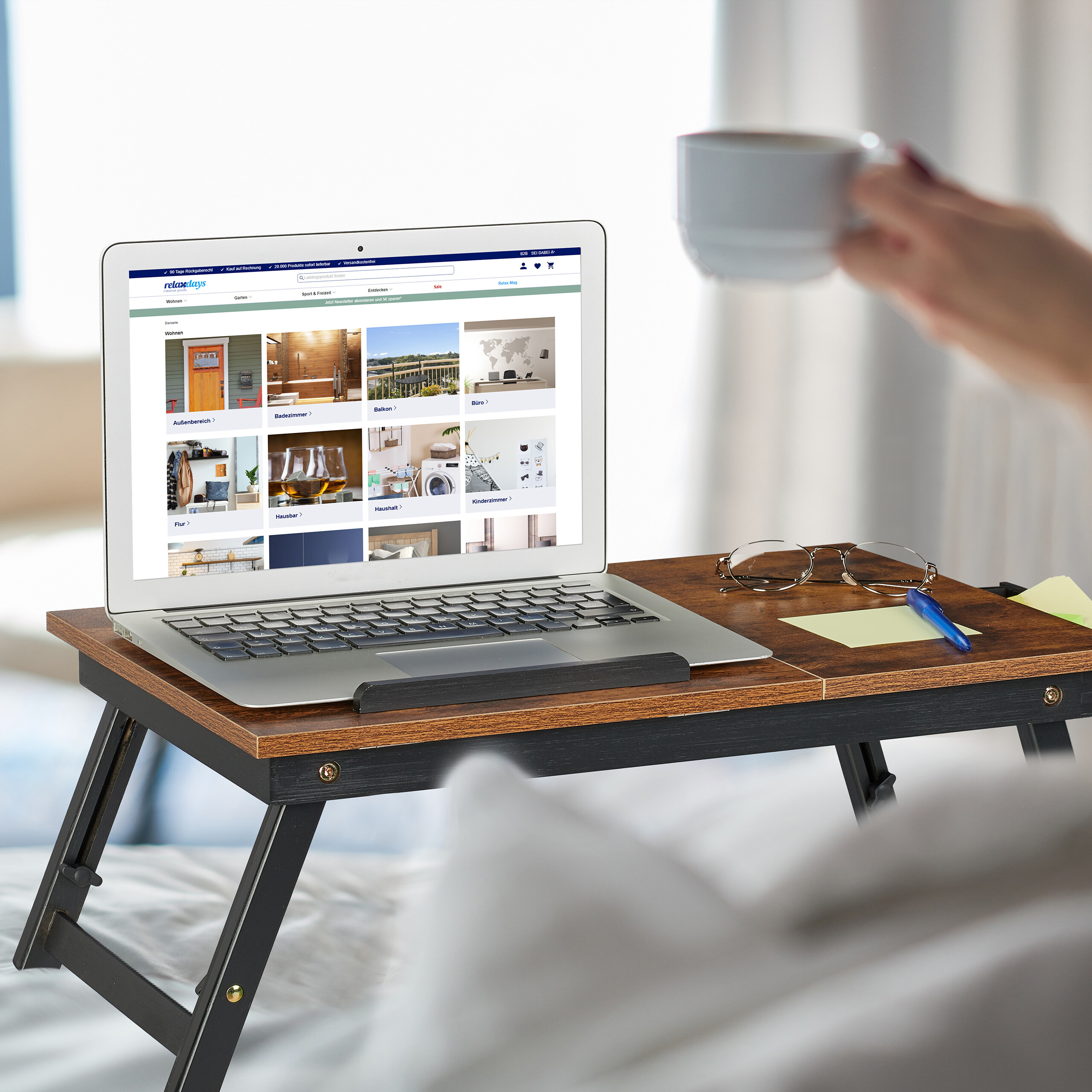 Ein Laptop auf einem Holztisch im Bett zeigt einen Online-Shop; daneben Brille, Stift und Tasse.