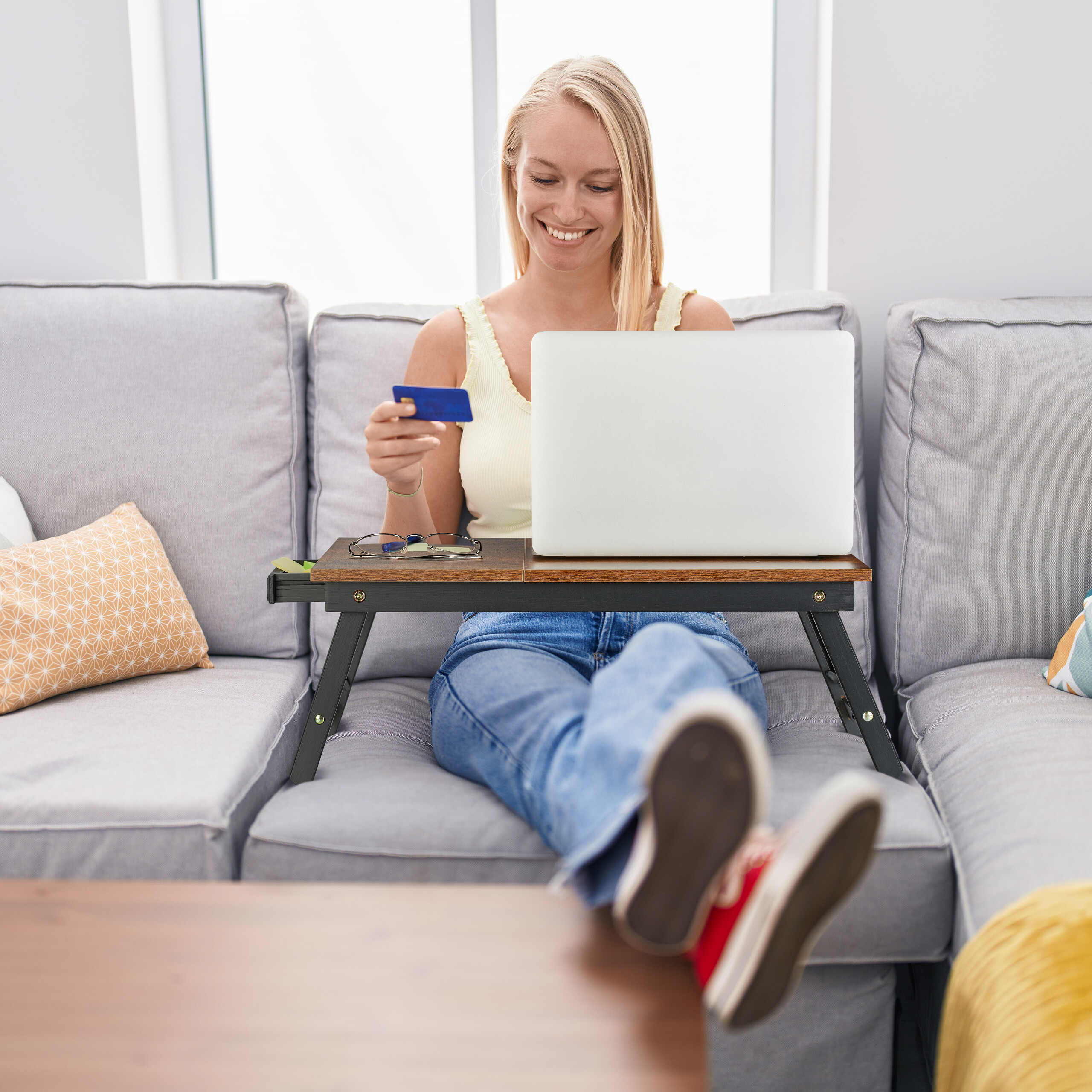 Blonde Frau sitzt auf einem Sofa, hält eine Kreditkarte und arbeitet am Laptop mit einem Schreibtischregal.