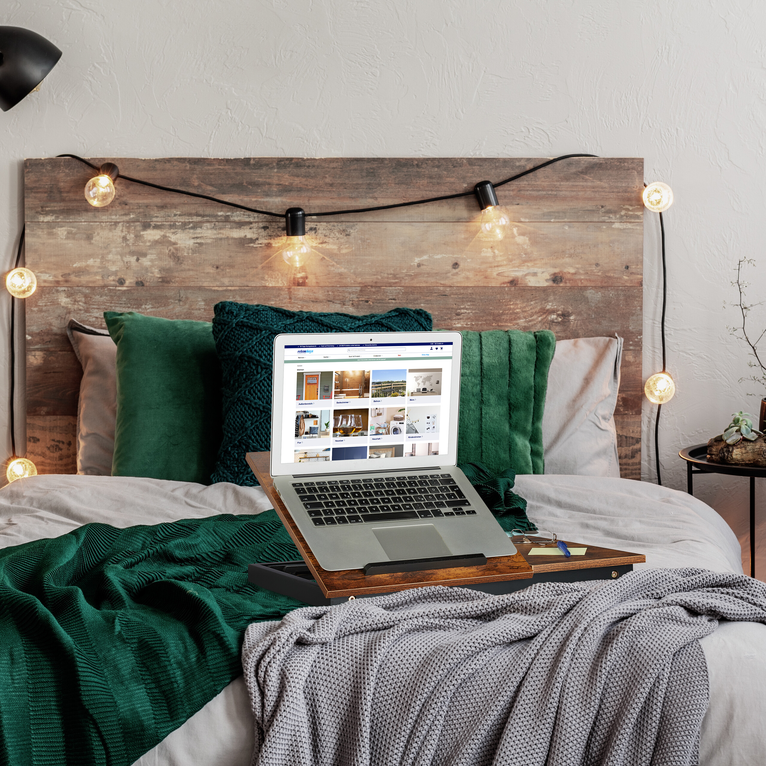 Ein Laptop auf einem Holztisch im Bett mit grünen Kissen und Lichterketten an der Wand.