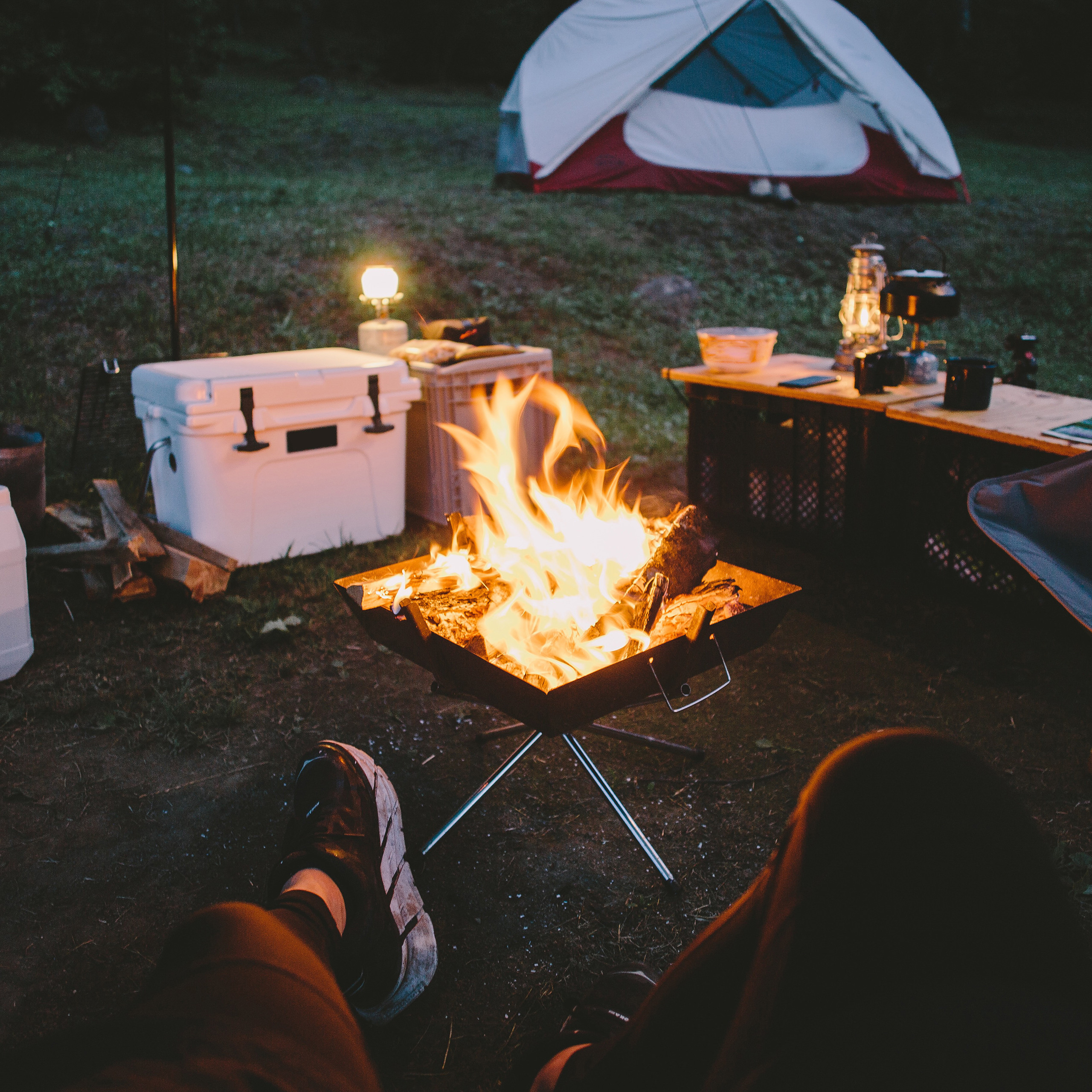 Ein Zeltplatz in der Dämmerung mit hellem Lagerfeuer, weißem Zelt, Kühlbox und Ausrüstung im Hintergrund.
