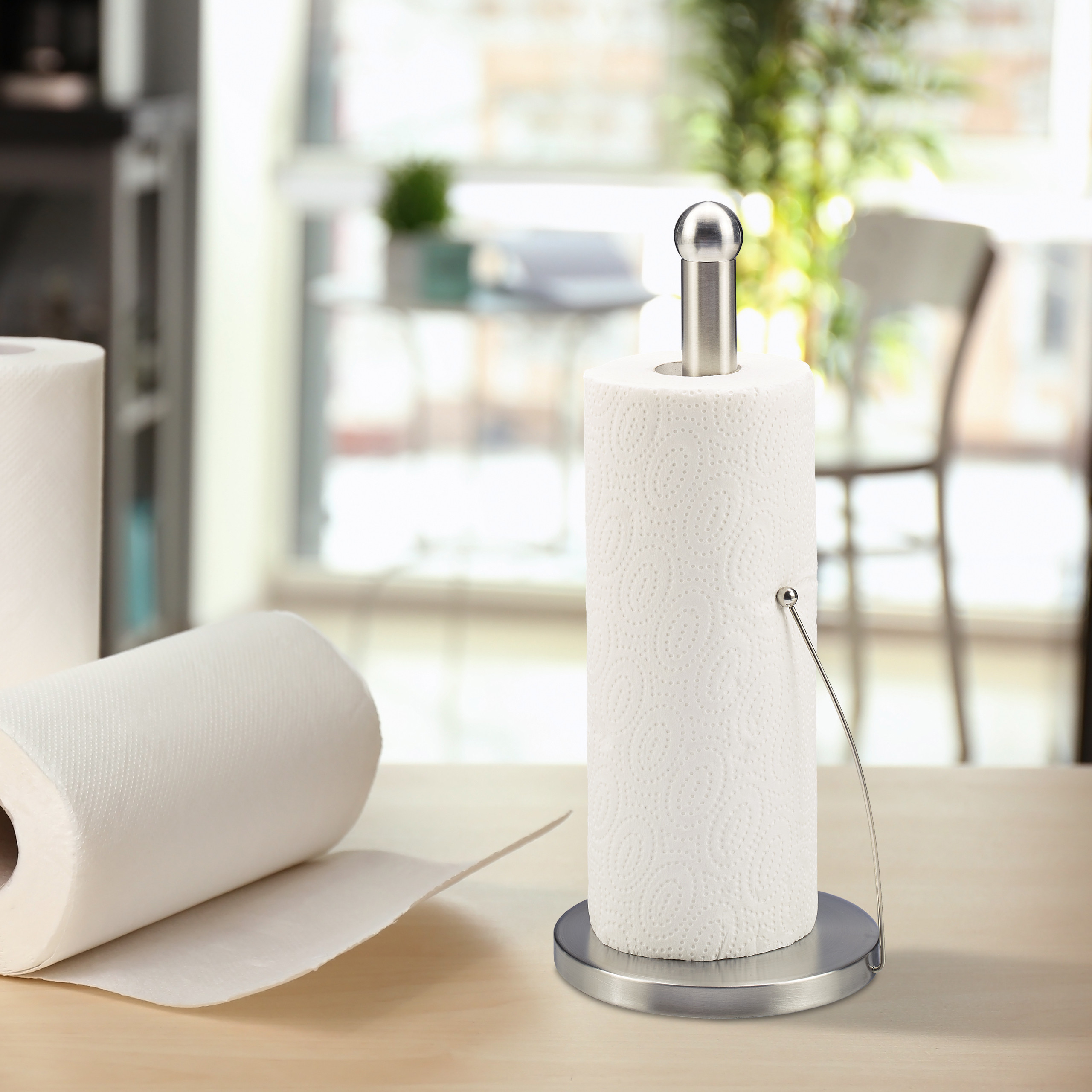 Ein stehender Küchenrollenhalter aus Edelstahl mit einer Papierrolle auf einem Holztisch, im Hintergrund ein helles Zimmer.