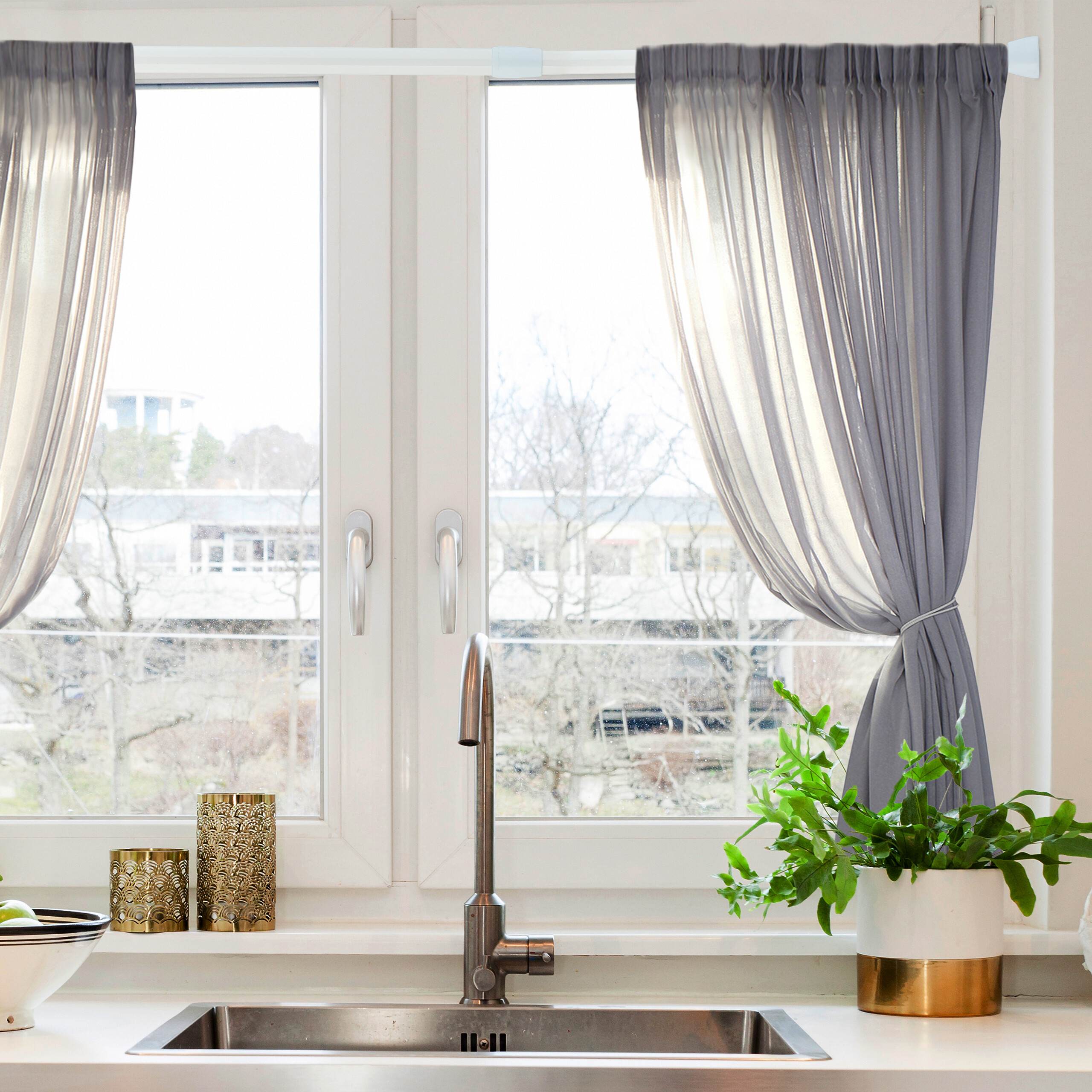 Eine Küchenspüle mit Wasserhahn vor einem Fenster mit grauen Vorhängen, goldenen Kerzenhaltern und einer Topfpflanze.