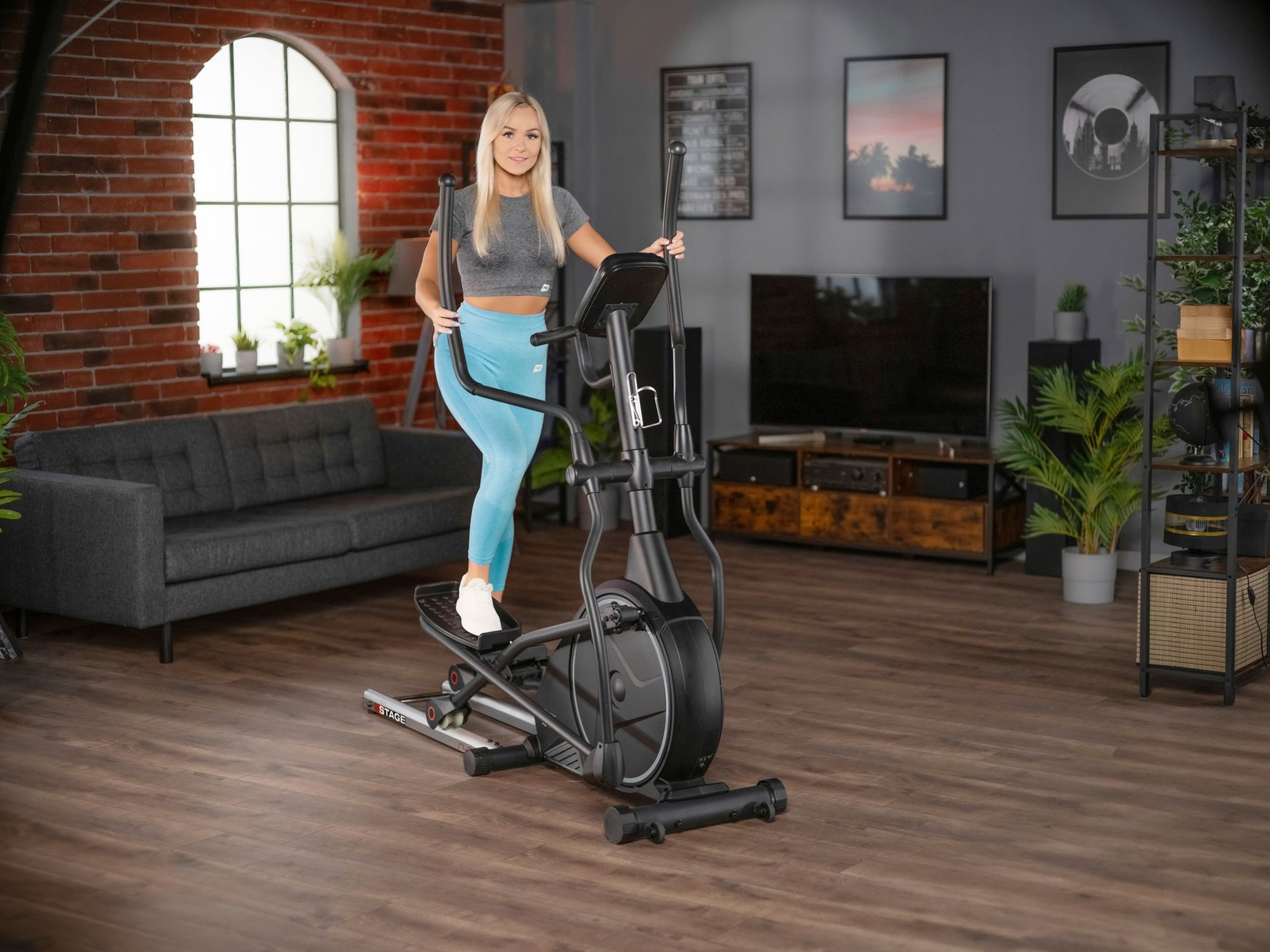 Eine blonde Frau im Sportoutfit trainiert zu Hause auf einem schwarzen Crosstrainer vor einer Backsteinwand.
