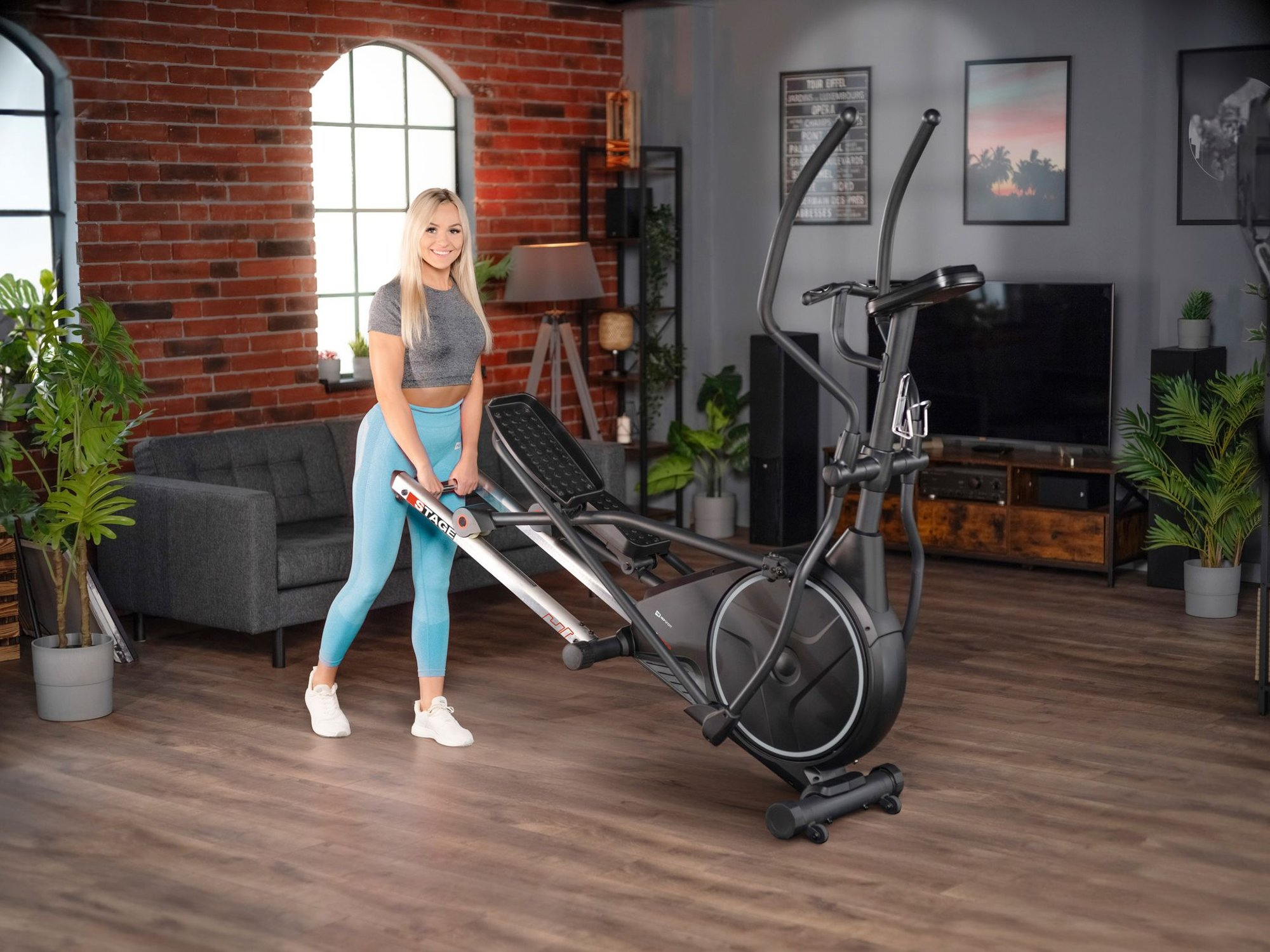 Eine blonde Frau in Sportkleidung lächelt und hält ein schwarzes Fitnessgerät in einem modernen Wohnzimmer.