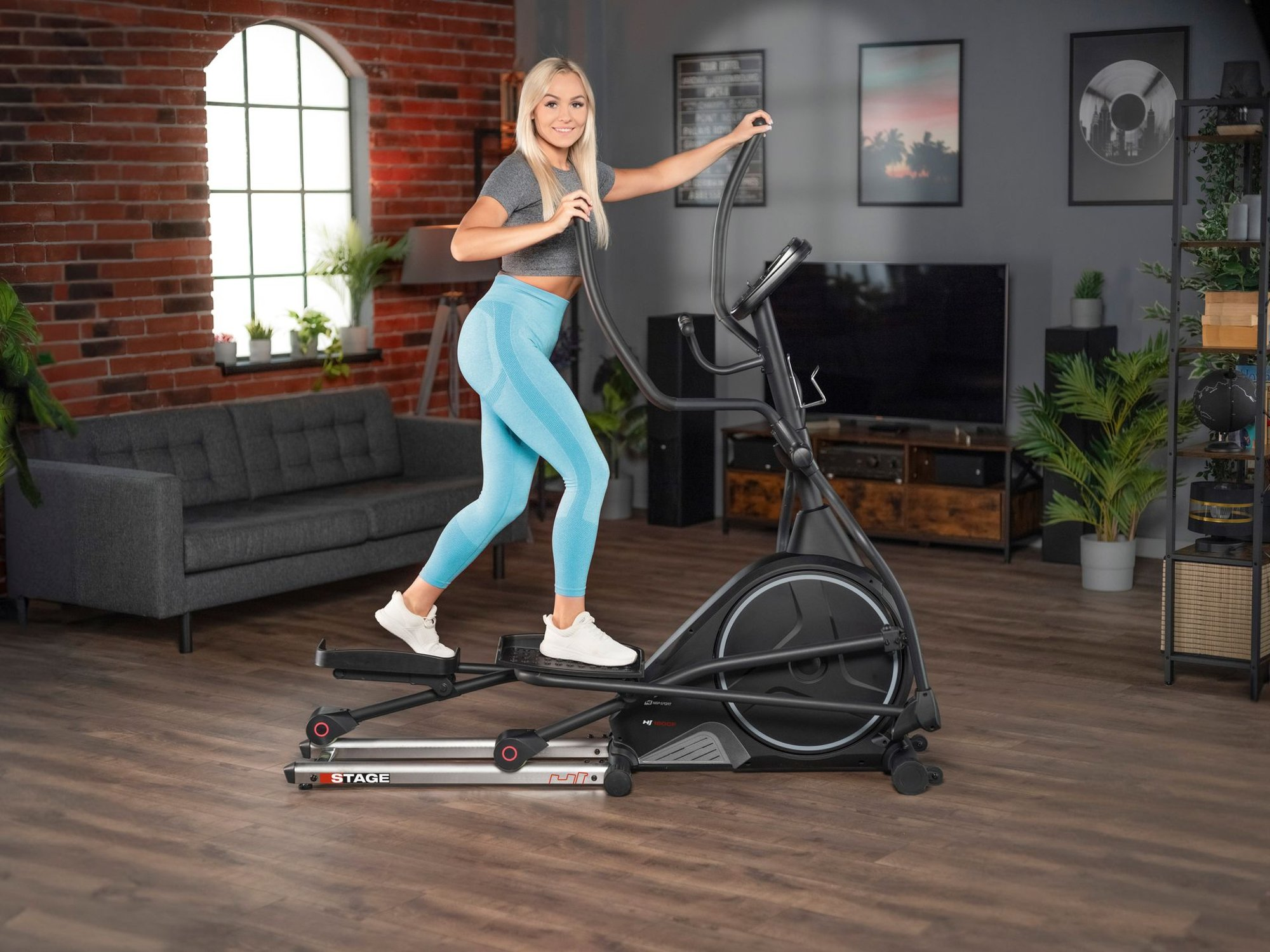 Eine blonde Frau im grauen Crop-Top und blauen Leggings trainiert auf einem schwarzen Crosstrainer im Wohnzimmer.
