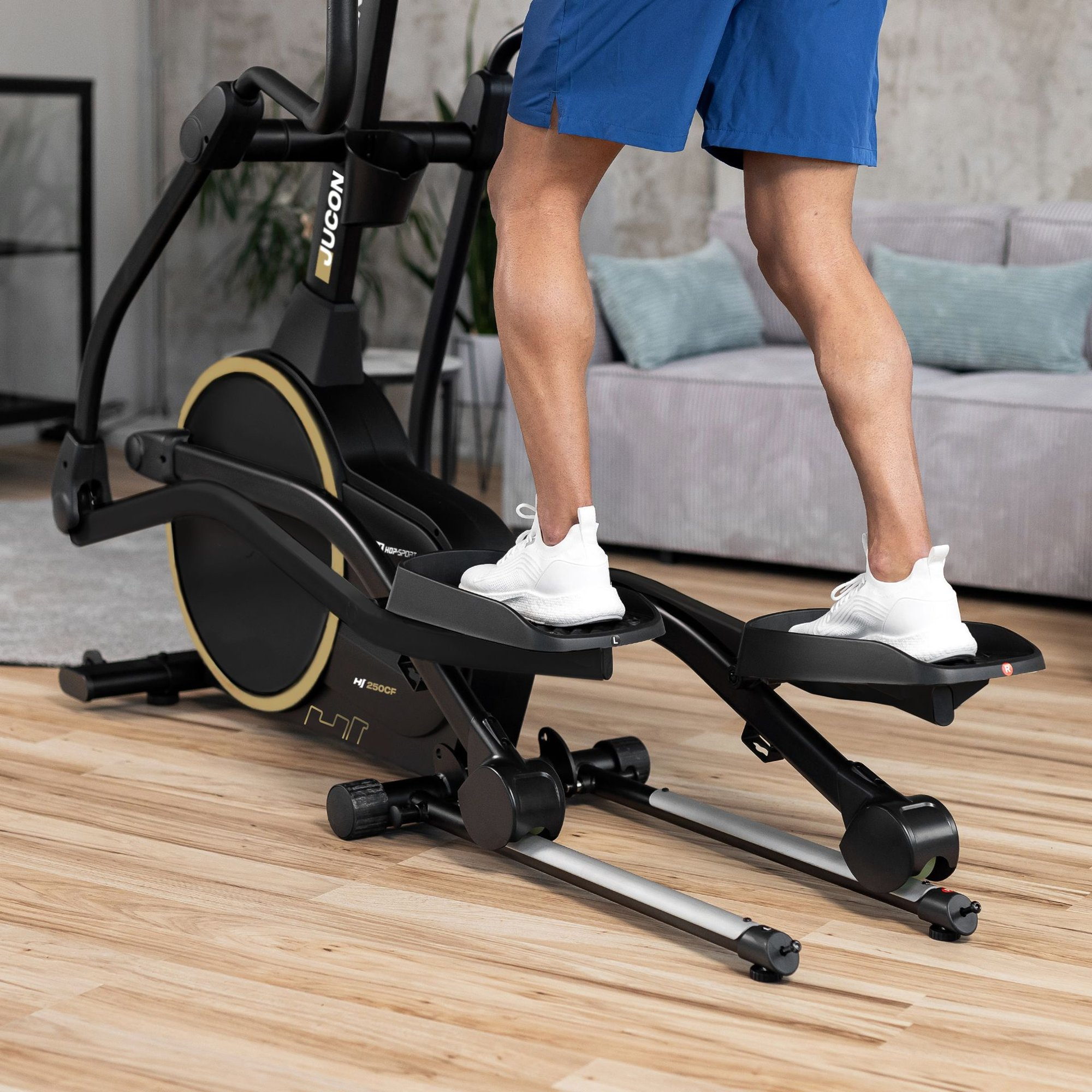 Person in blauen Shorts und weißen Sneakers trainiert zu Hause auf einem schwarzen Crosstrainer.
