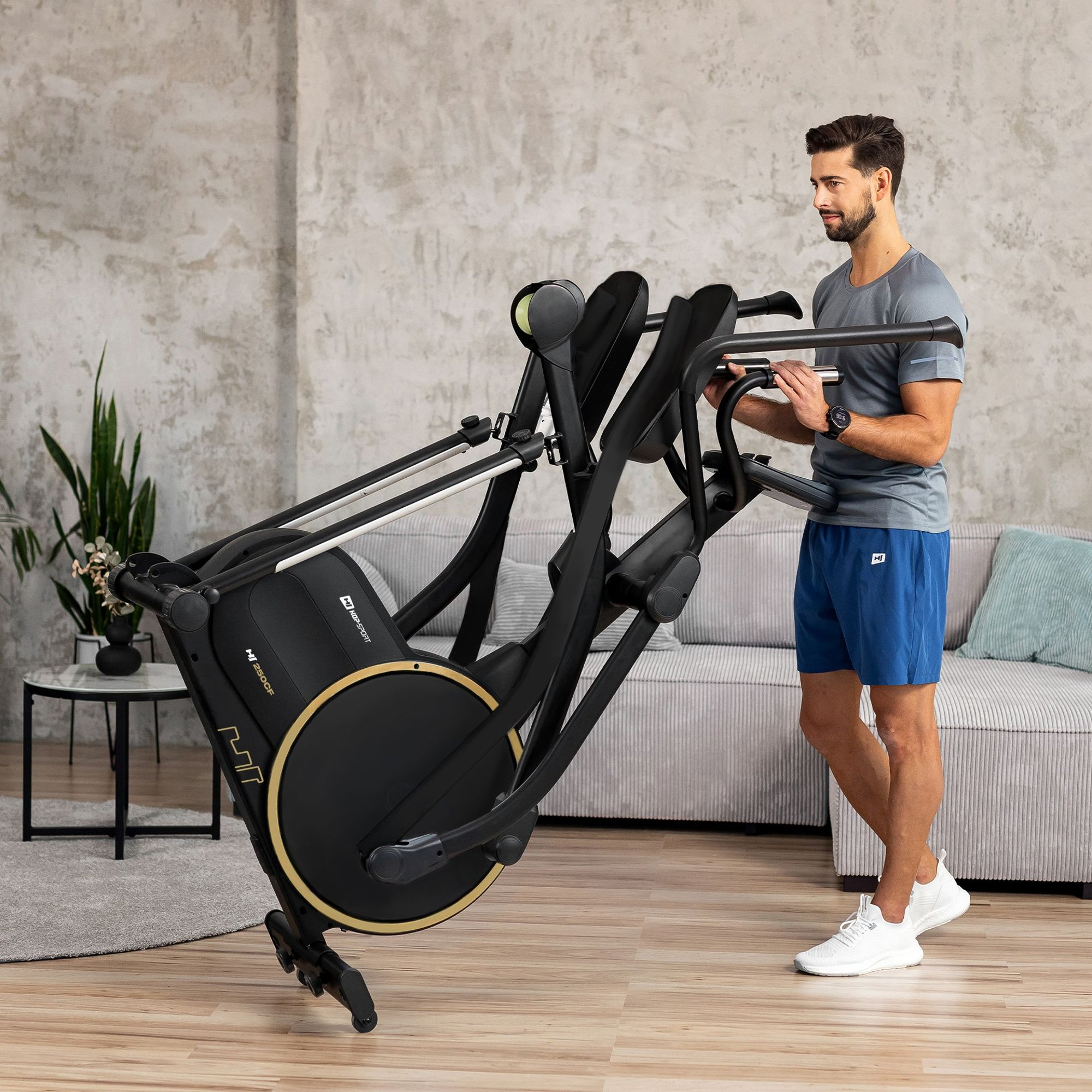 Ein Mann im grauen T-Shirt und blauen Shorts klappt in einem Wohnzimmer einen Crosstrainer zusammen.