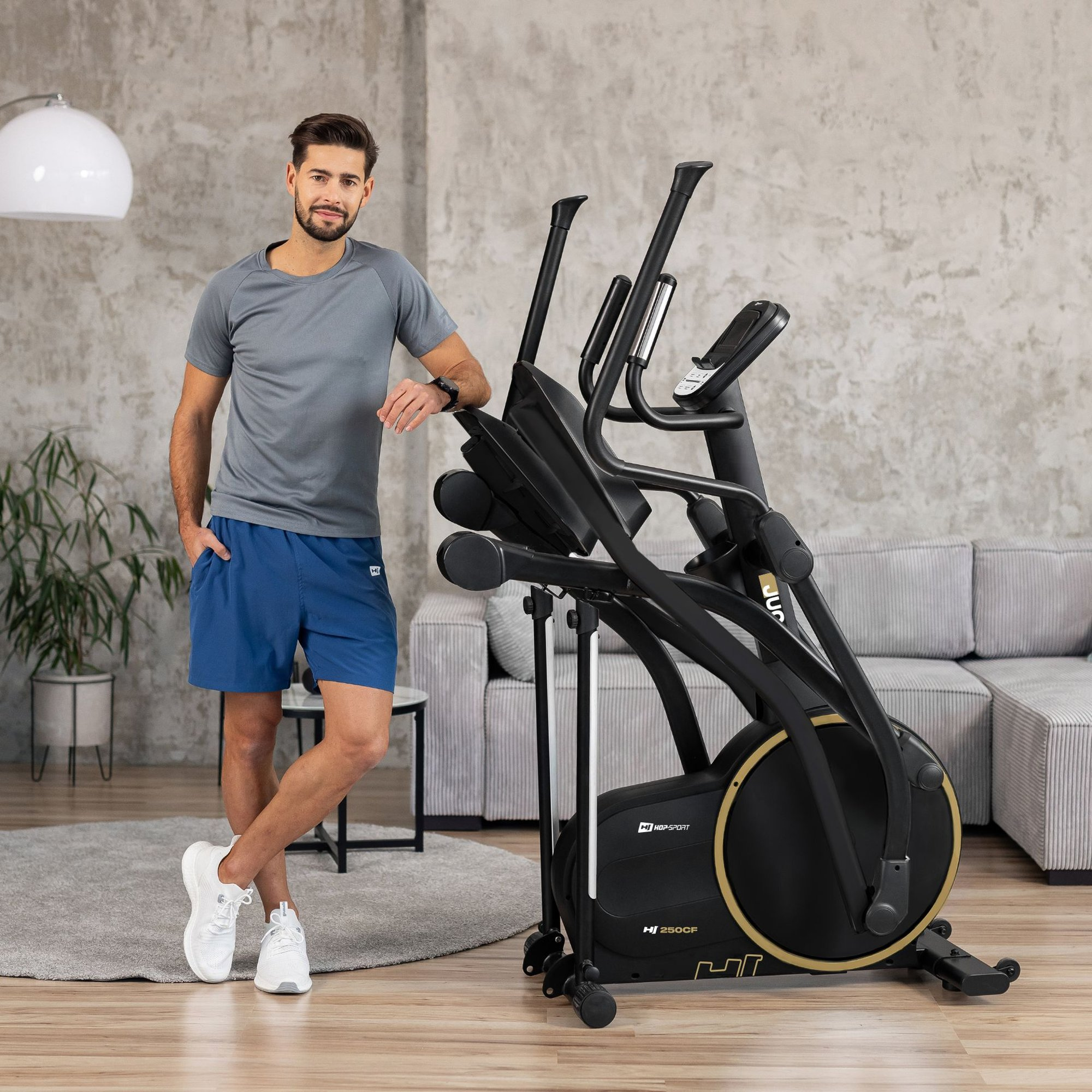 Ein lächelnder Mann in Sportkleidung lehnt sich an einen schwarzen Crosstrainer im Wohnzimmer.