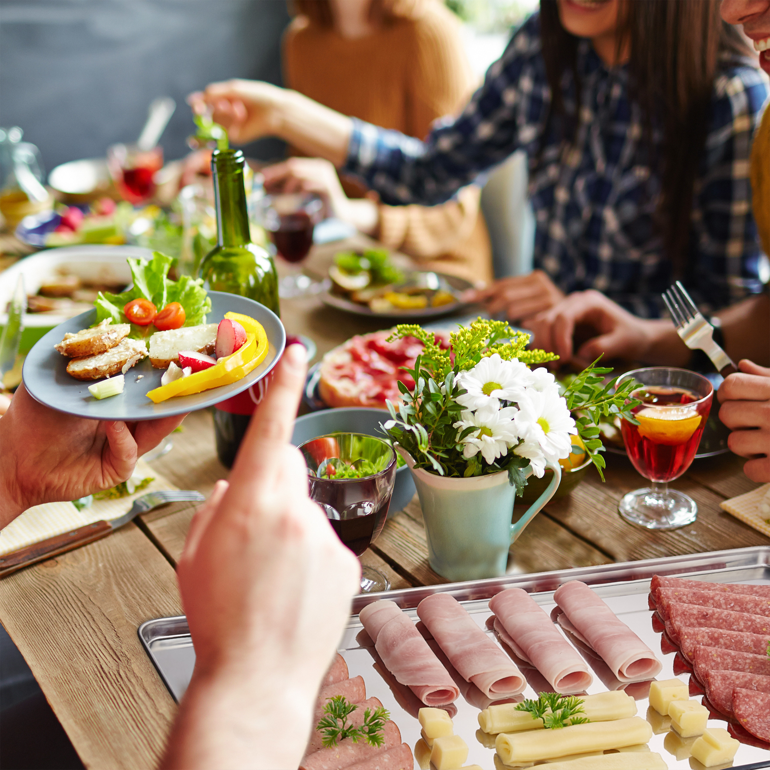 Freunde essen zusammen an einem Tisch mit Salat, Aufschnitt und Blumen.