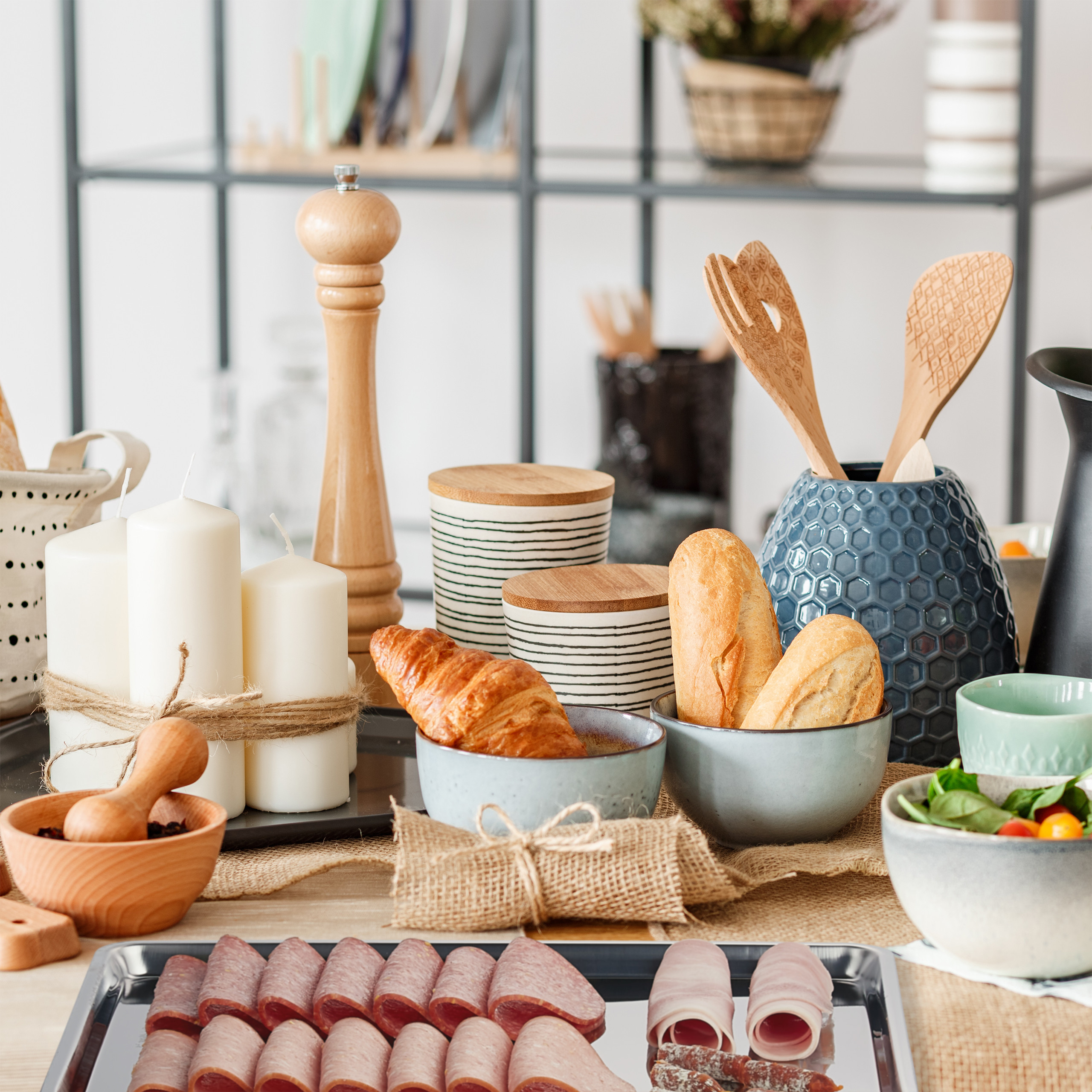 Frühstückstisch mit Brot, Croissant, Aufschnitt, Salat und Kerzen in einer hellen Küche.