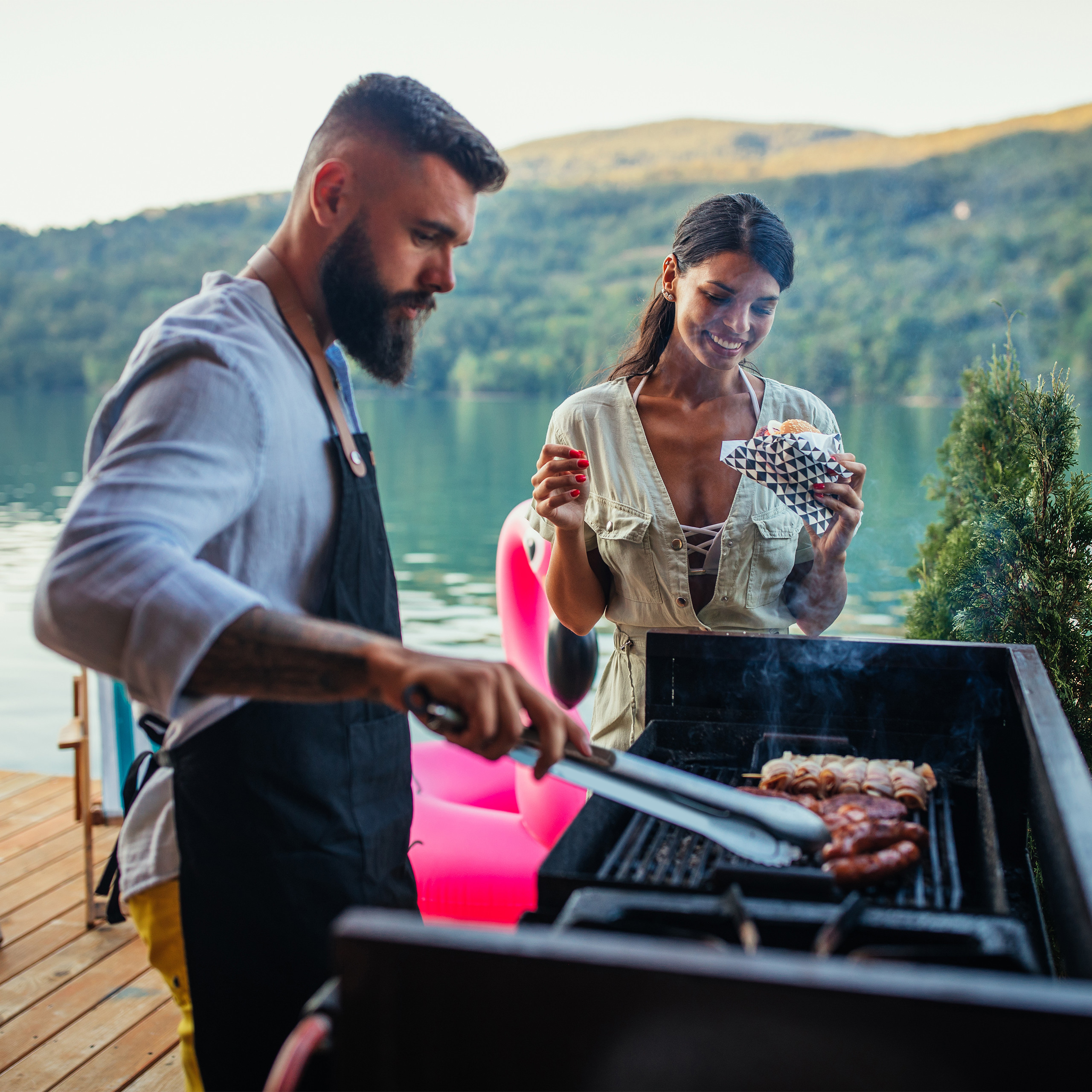 Ein bärtiger Mann grillt Fleisch, während eine Frau daneben steht und einen Burger isst, an einem See.
