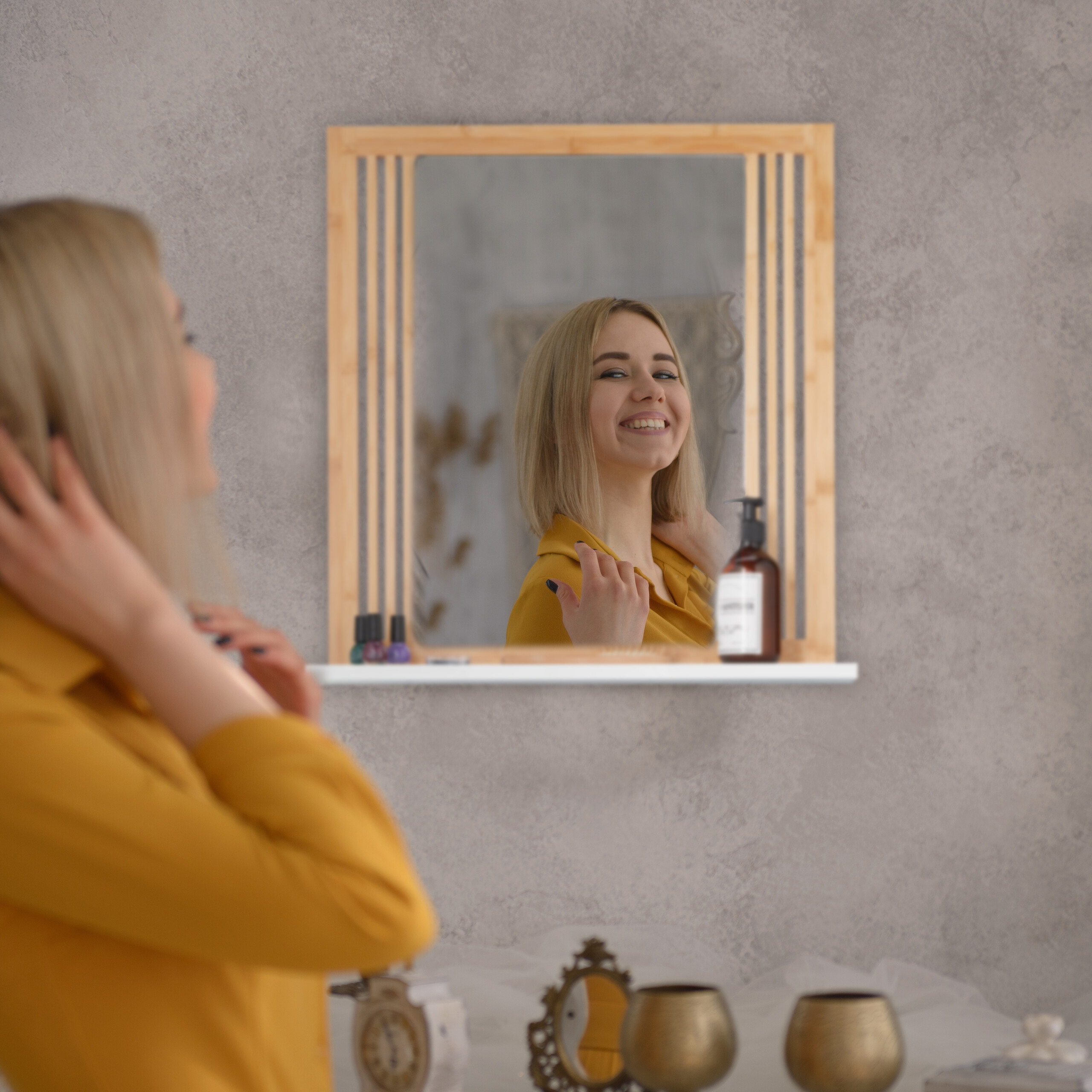 Eine blonde Frau im gelben Hemd lächelt in einen Spiegel mit Holzrahmen.