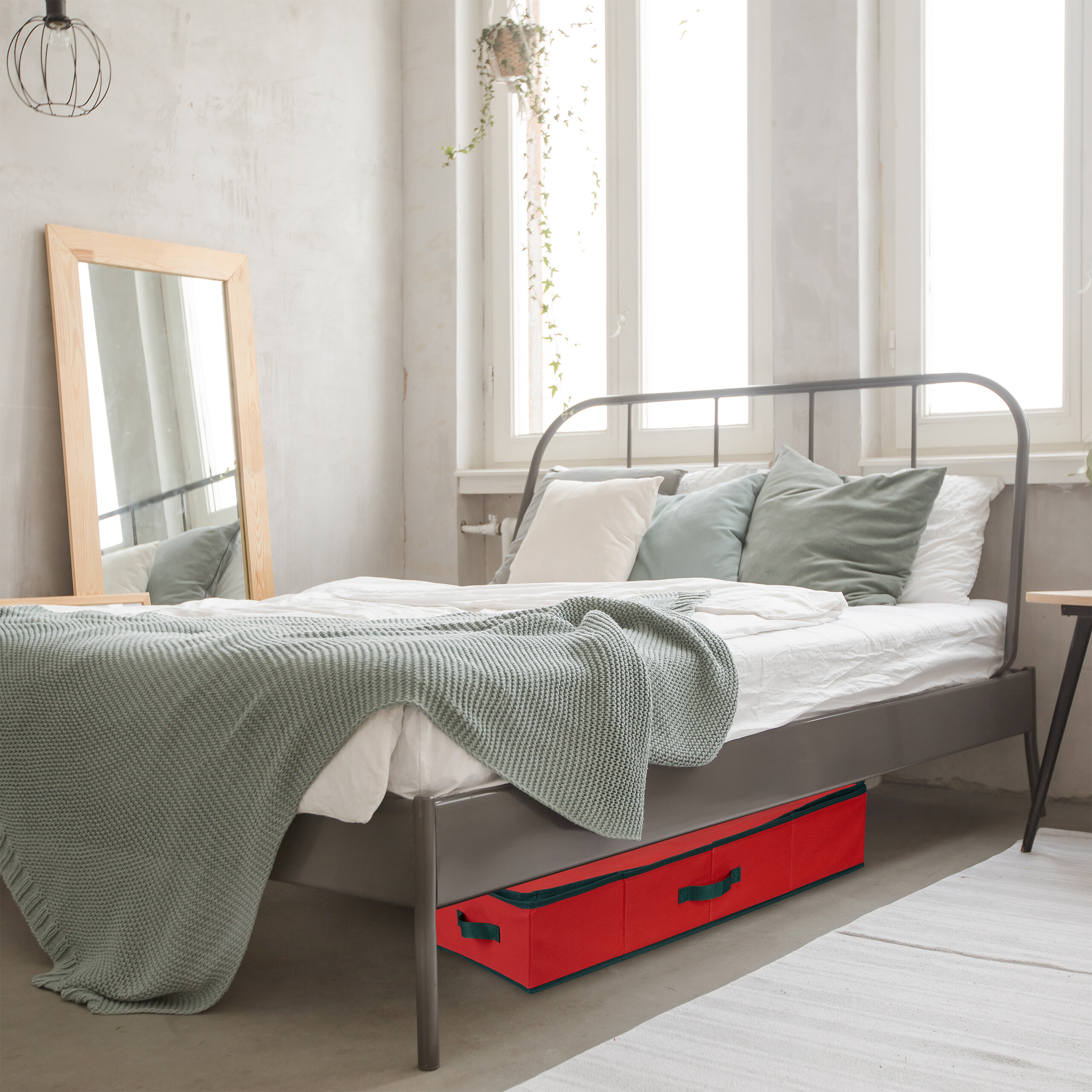 Ein graues Bett mit Kissen und grüner Decke in einem hellen Zimmer, eine rote Aufbewahrungsbox darunter.