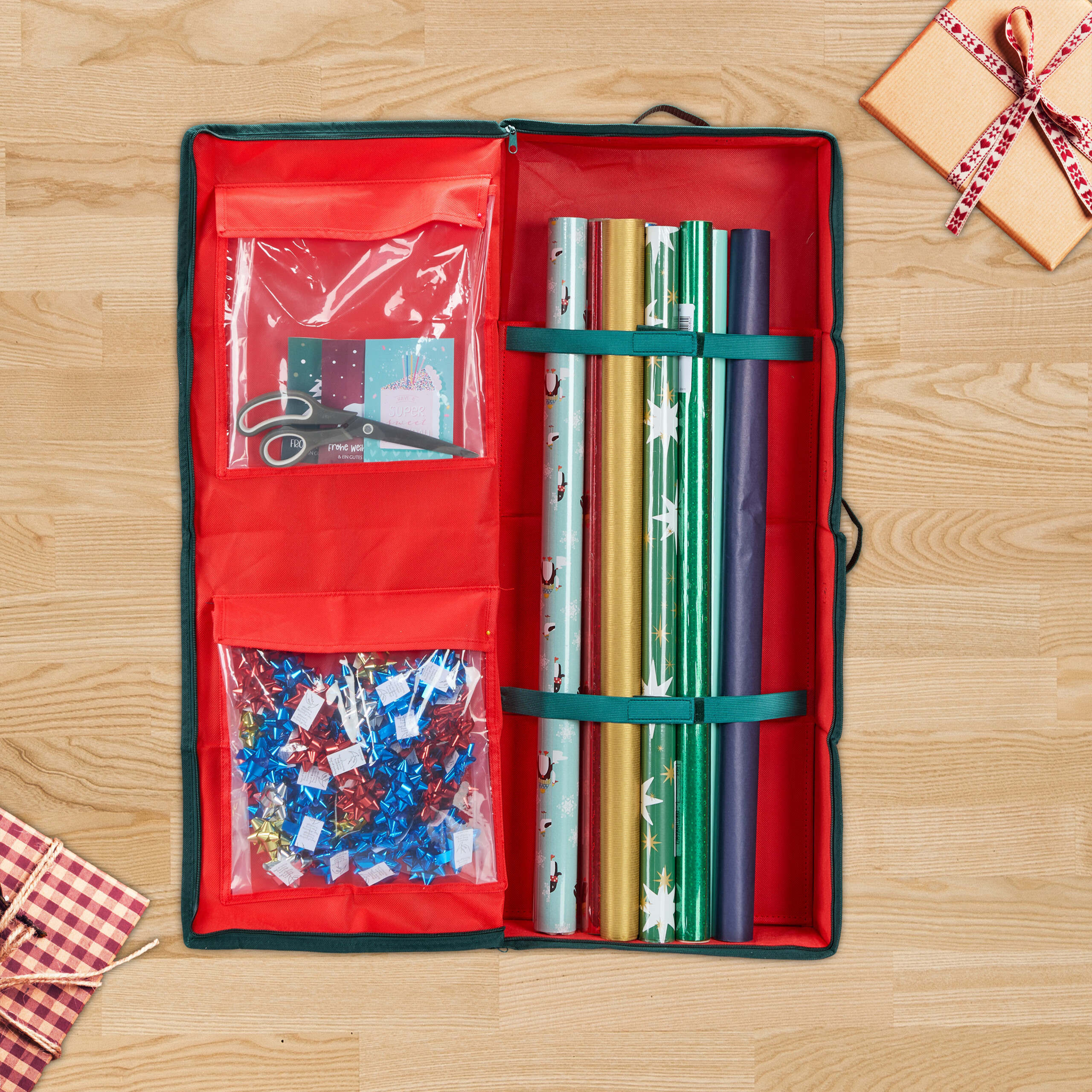 Ein offener roter Geschenkpapier-Organizer auf Holz, gefüllt mit Rollen, Schleifen und einer Schere.