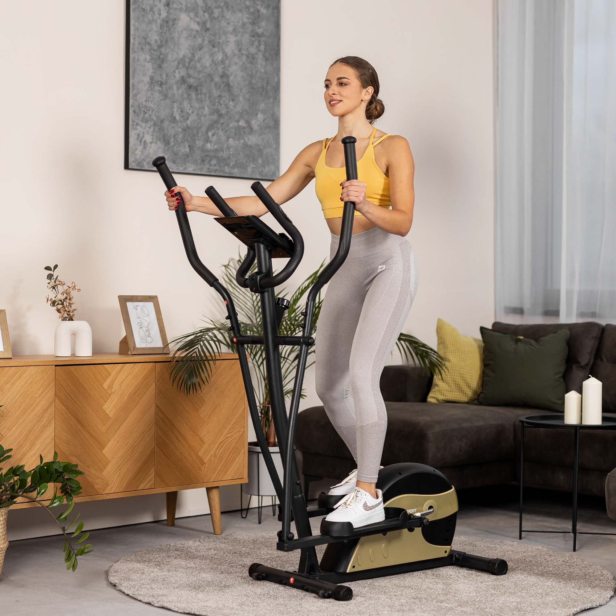 Eine Frau in gelbem Oberteil und grauen Leggings trainiert in einem hellen, modernen Wohnzimmer auf einem Crosstrainer.