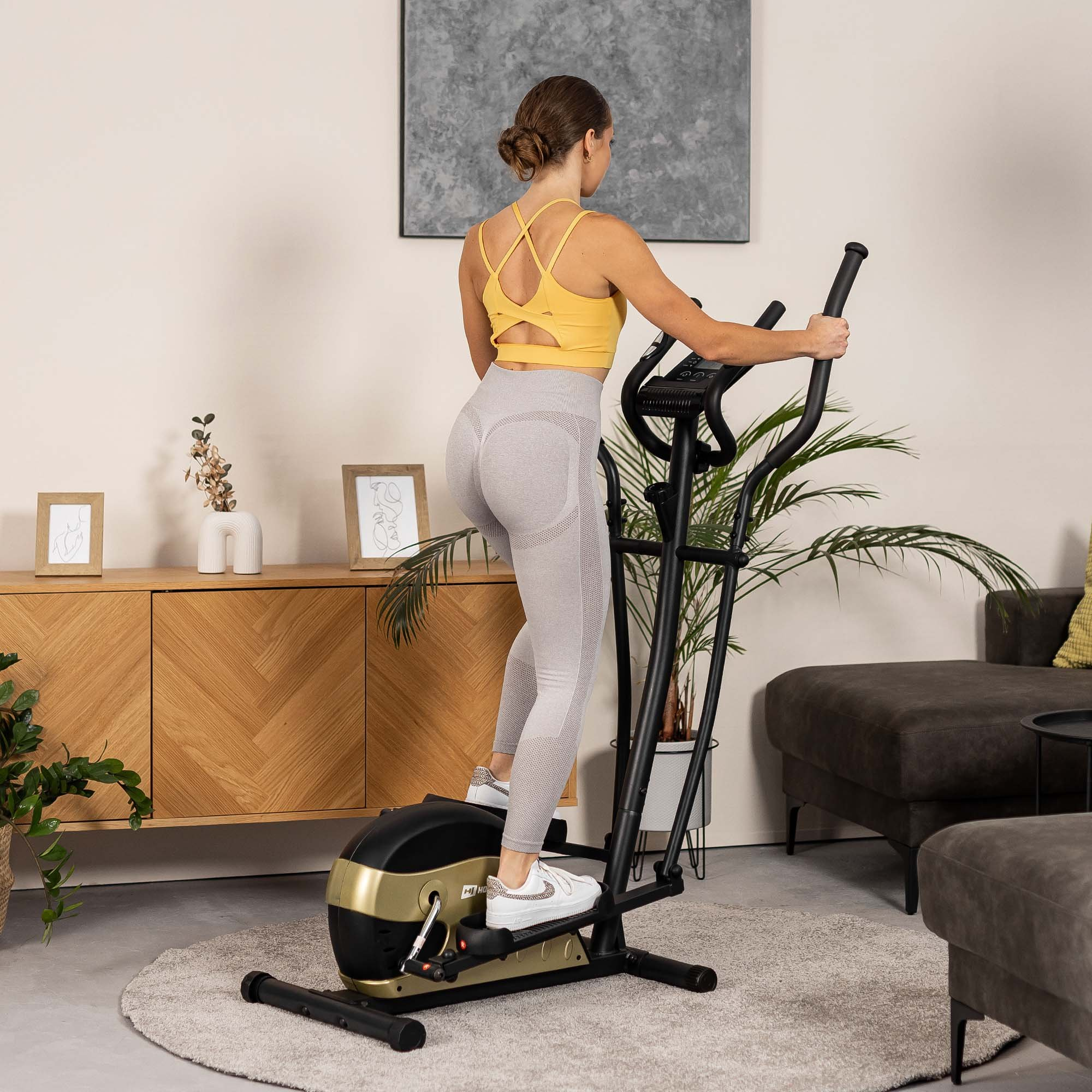Eine Frau in gelbem Oberteil und grauen Leggings trainiert in einem hellen Wohnzimmer auf einem schwarz-goldenen Crosstrainer.