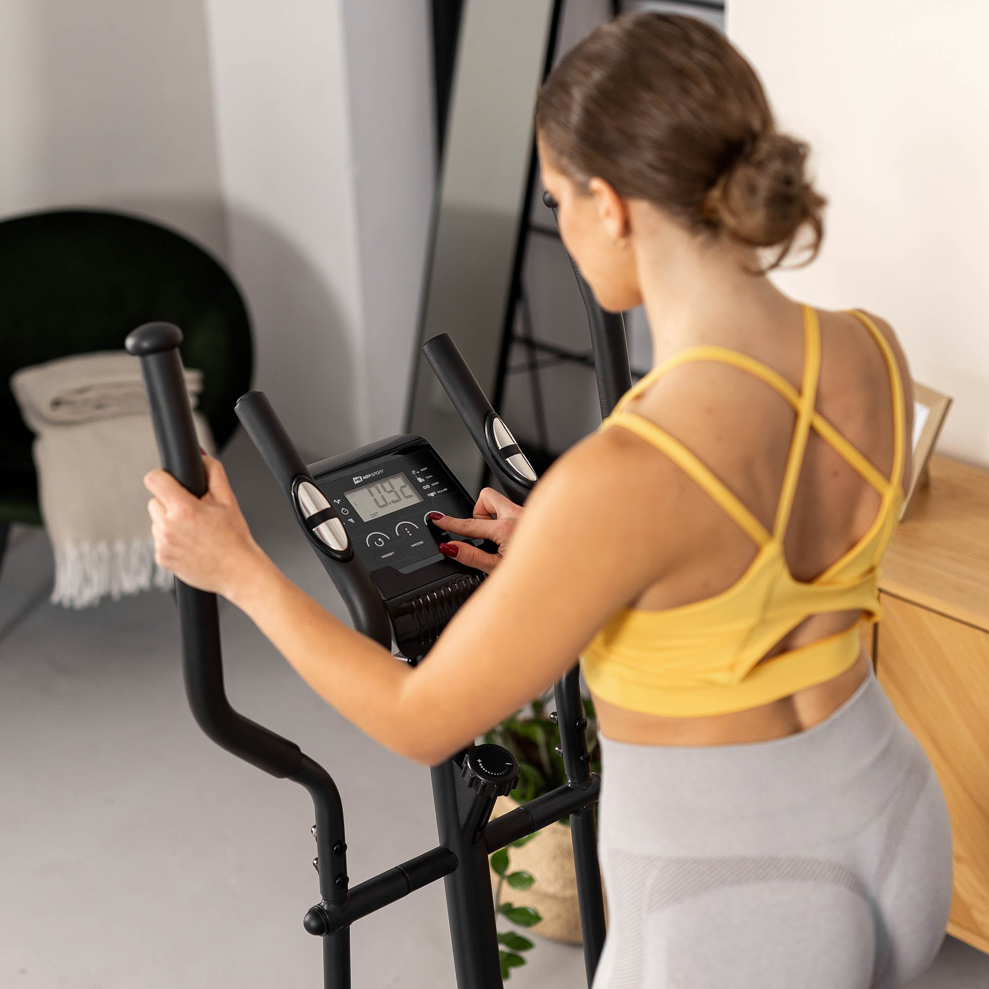 Eine Frau im gelben Sport-Top trainiert drinnen auf einem Heimtrainer und drückt Knöpfe.