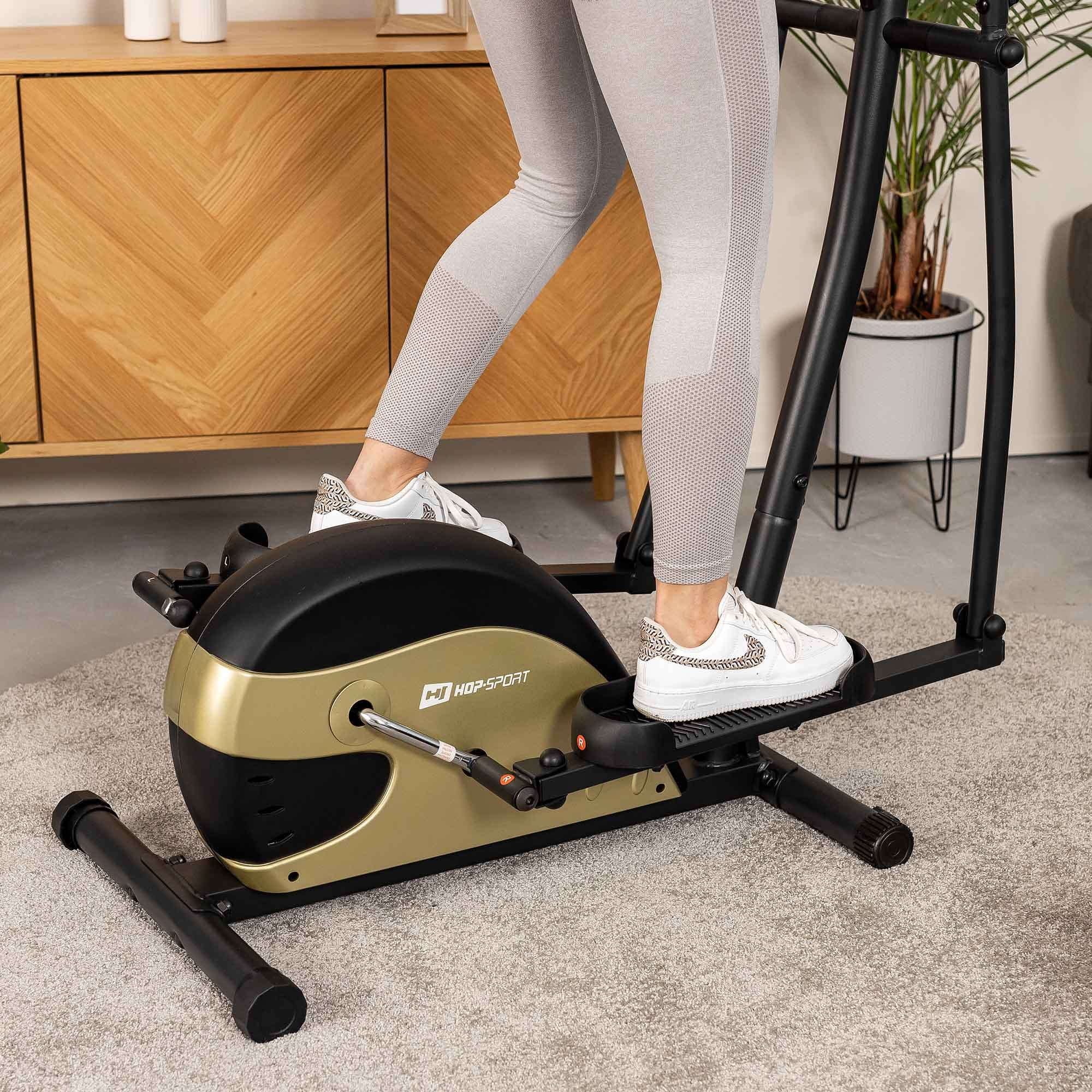 Person in grauen Leggings trainiert auf einem schwarz-goldenen Crosstrainer im Wohnzimmer.