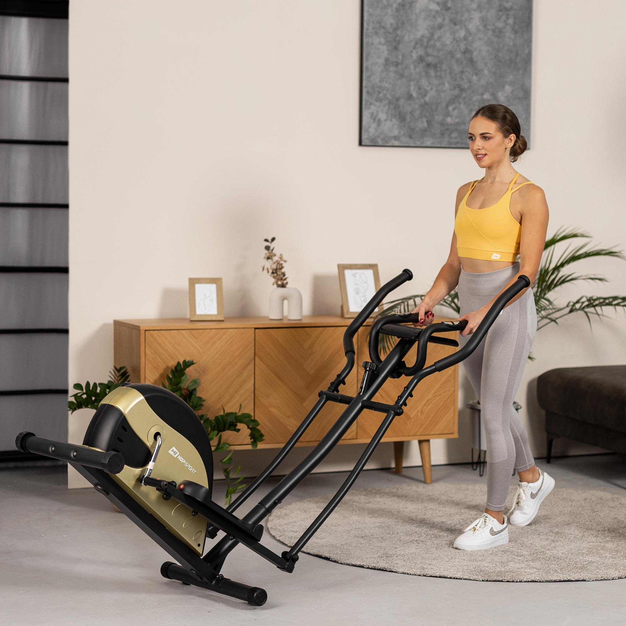 Eine Frau im gelben Oberteil und grauen Leggings trainiert in einem hellen Raum auf einem Crosstrainer.