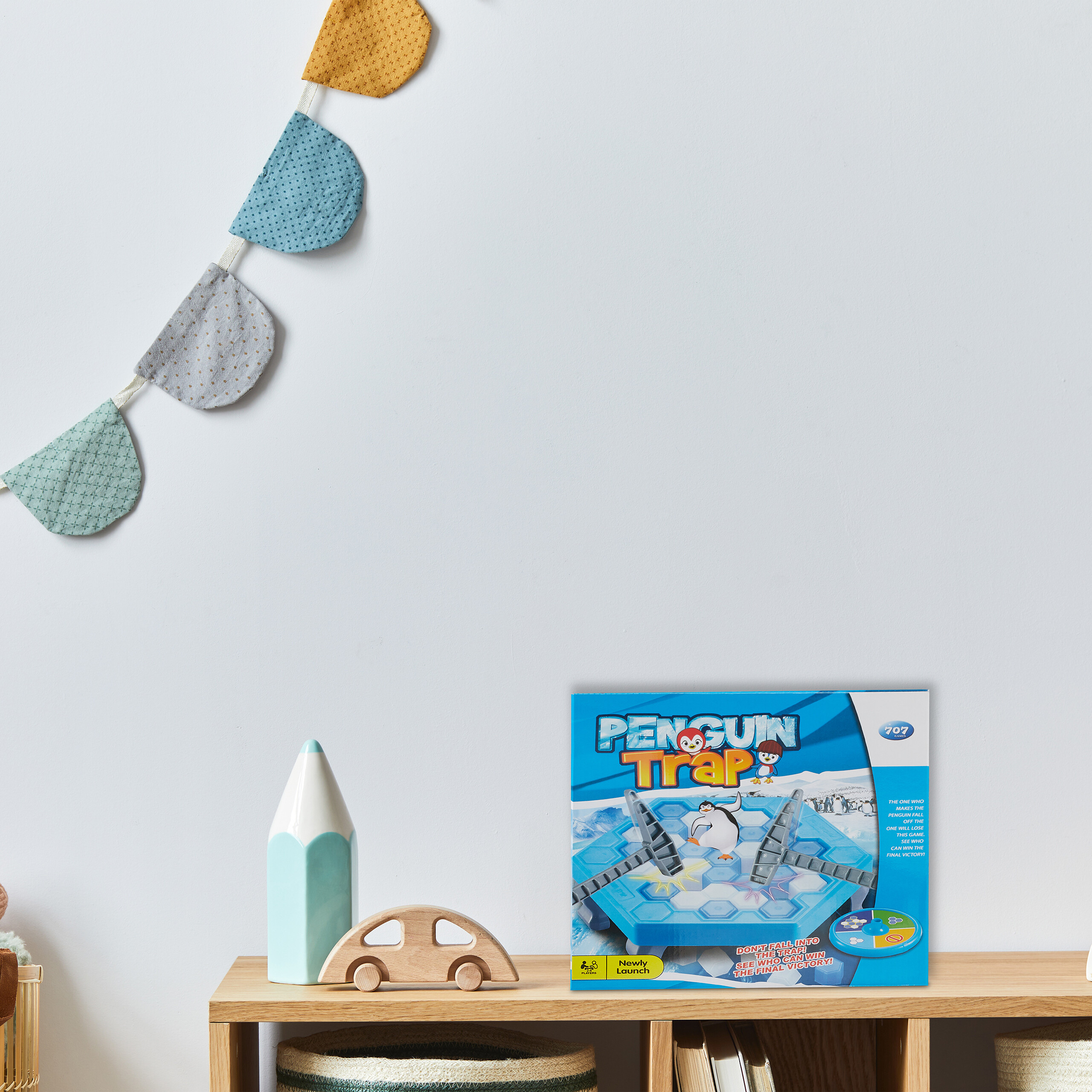 Ein Holzregal mit Pinguin-Falle-Spiel, Spielzeugauto und Bleistift-Dekoration unter einer bunten Stoffgirlande.