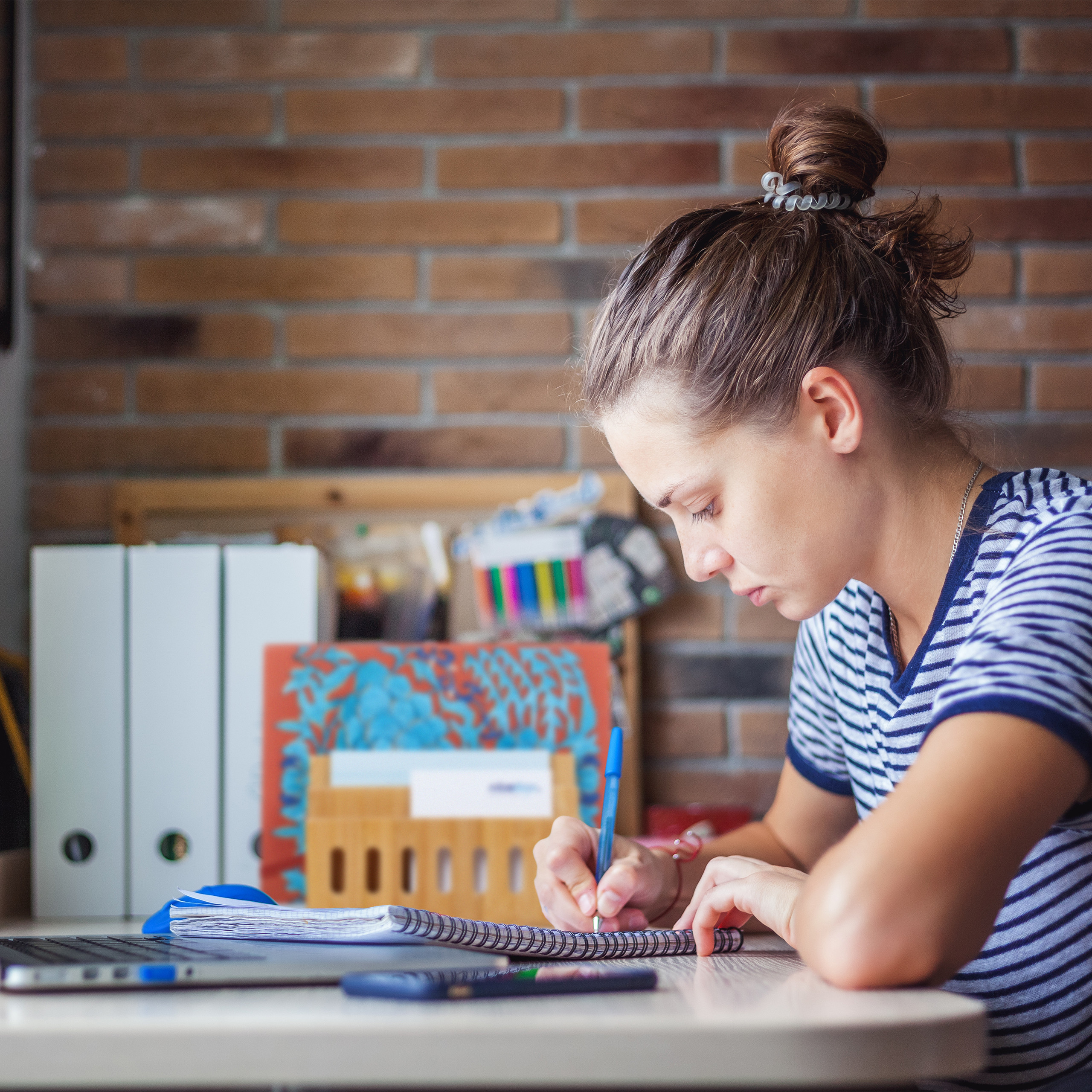 Eine Frau in einem gestreiften Shirt schreibt an einem Schreibtisch mit Laptop und Ordnern.