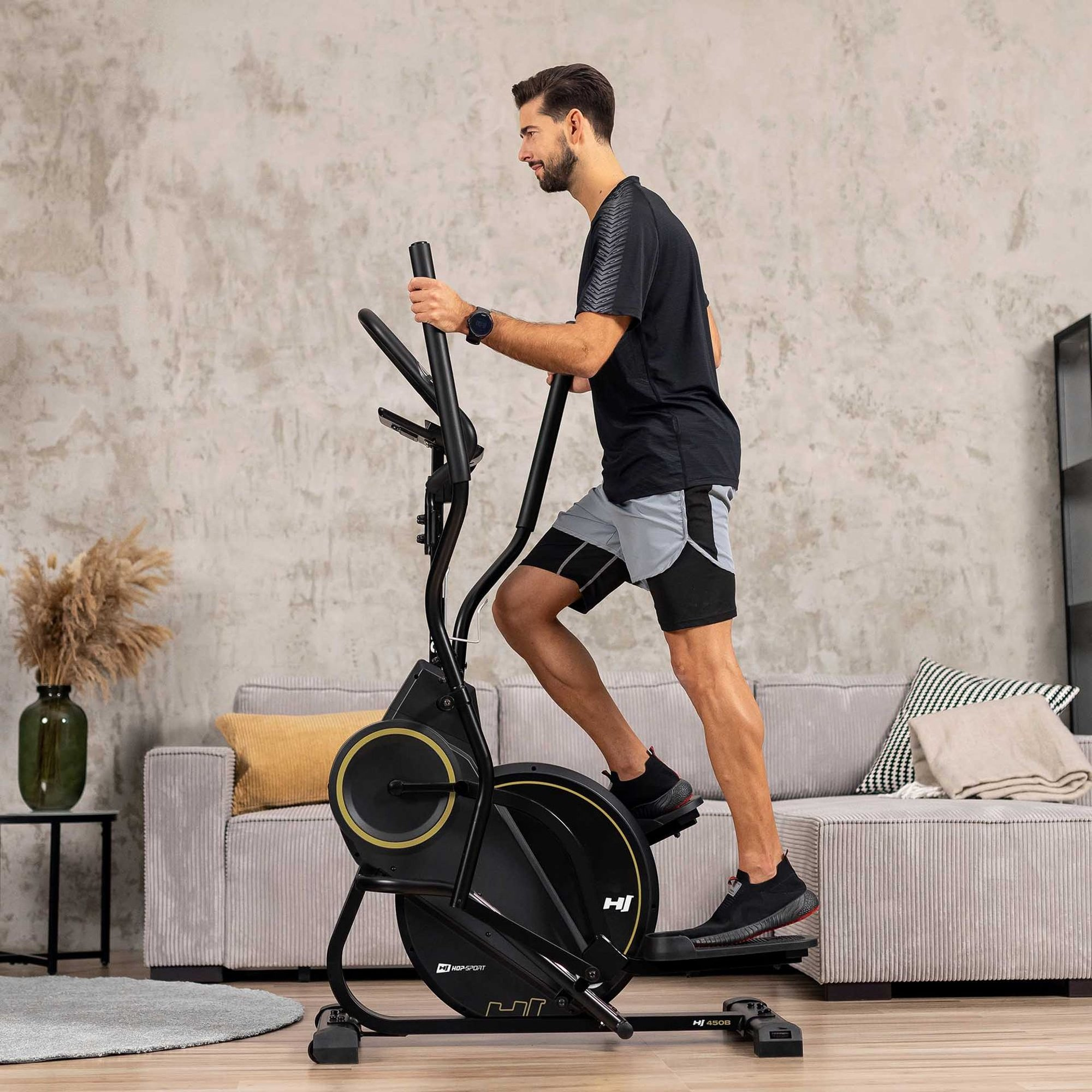 Mann trainiert auf einem schwarzen Crosstrainer in einem modernen Wohnzimmer mit Sofa und Pflanzen.