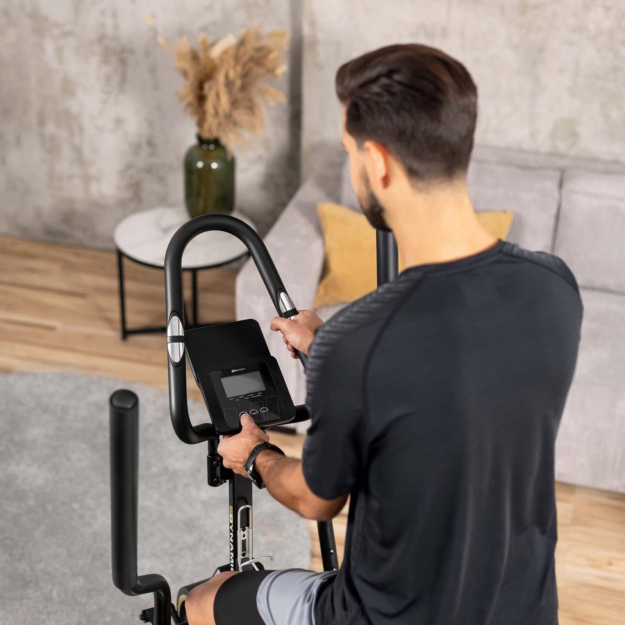 Ein Mann in Sportkleidung trainiert zu Hause auf einem Heimtrainer vor einer grauen Wand.