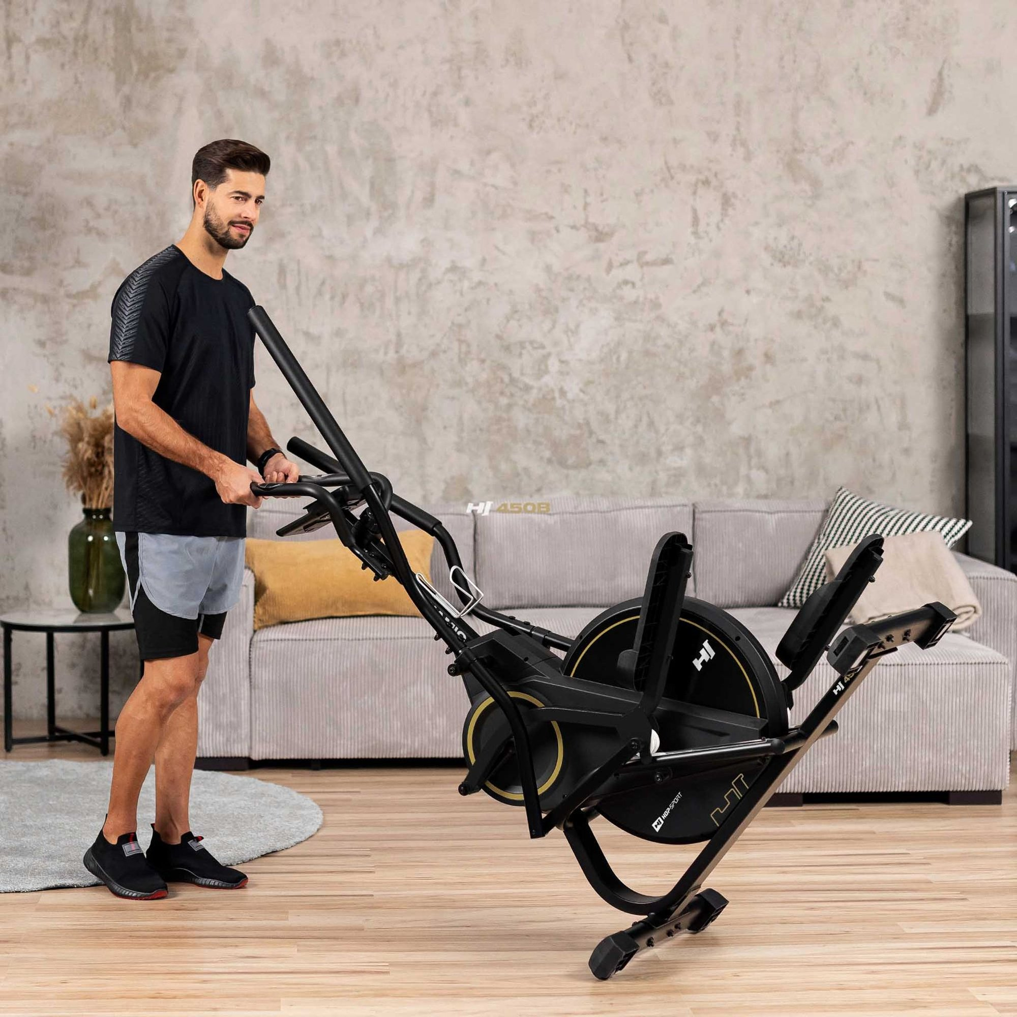 Ein Mann im schwarzen T-Shirt und grauen Shorts transportiert einen Heimtrainer in einem modernen Wohnzimmer.