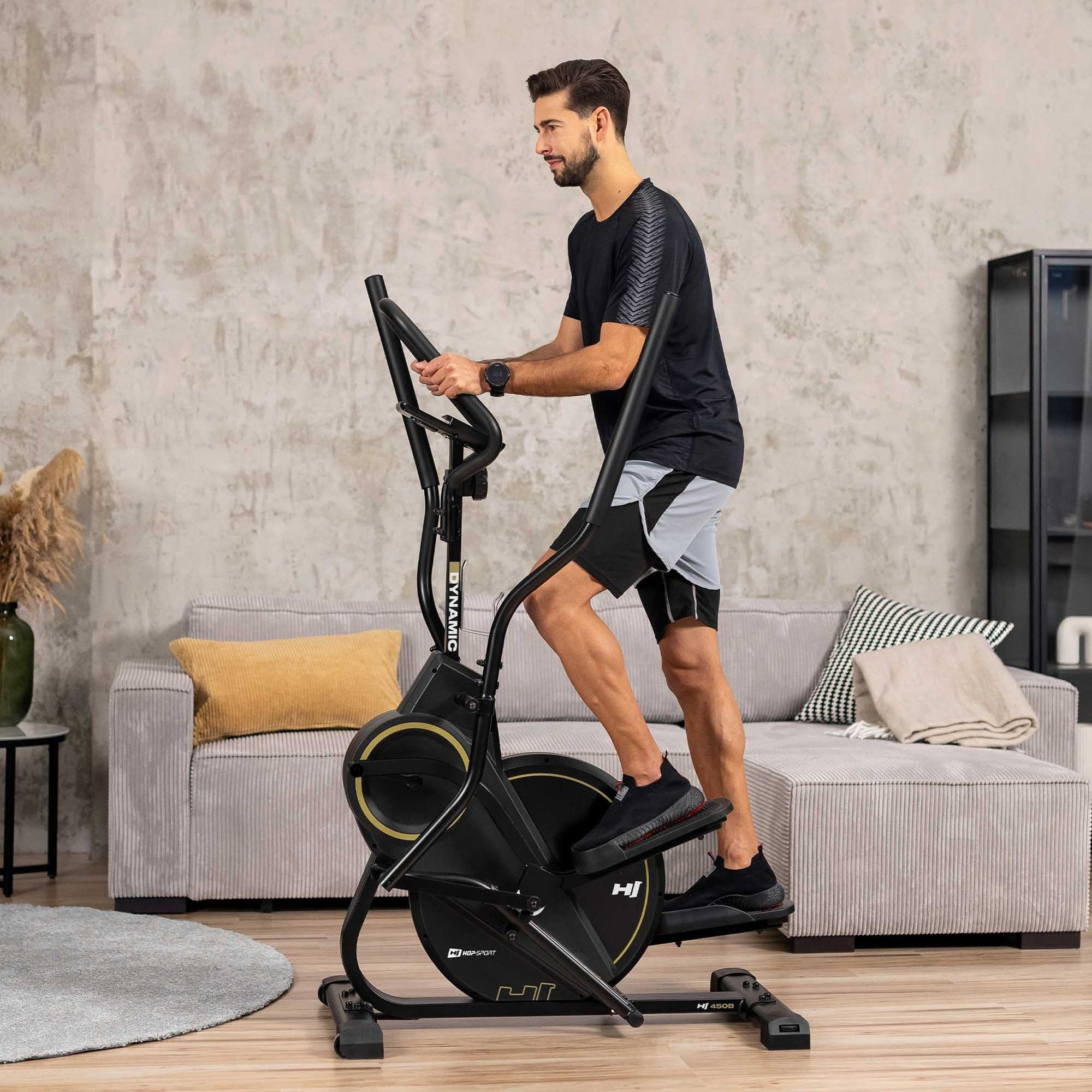 Ein Mann in Sportkleidung trainiert zu Hause auf einem schwarzen Crosstrainer vor einem Sofa.
