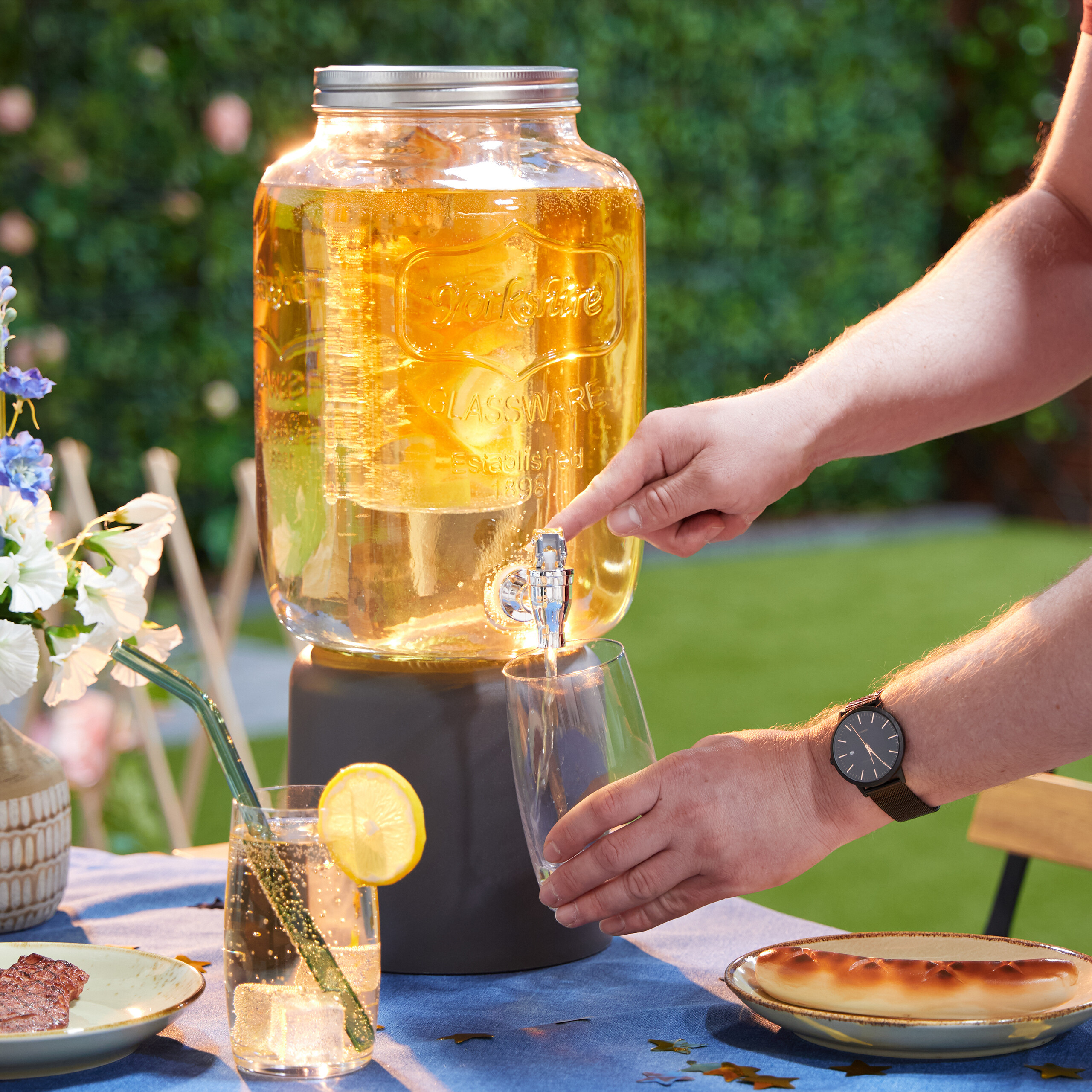 Eine Person gießt ein Getränk aus einem großen Glasbehälter in ein Glas auf einem Tisch.