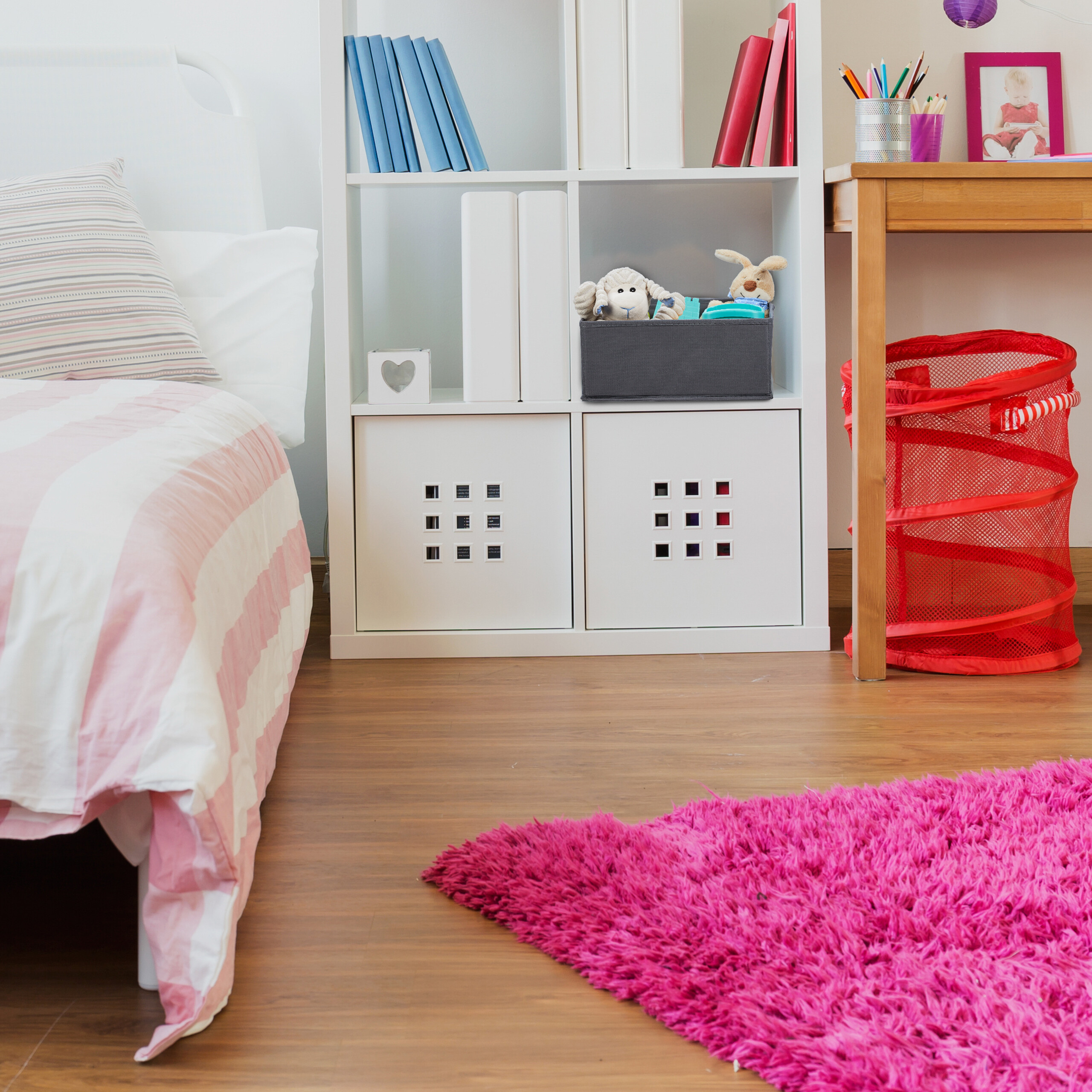 Ein kinderzimmer mit weißem regal, rosafarbenem bett, holztisch, rotem korb und pinkem teppich.
