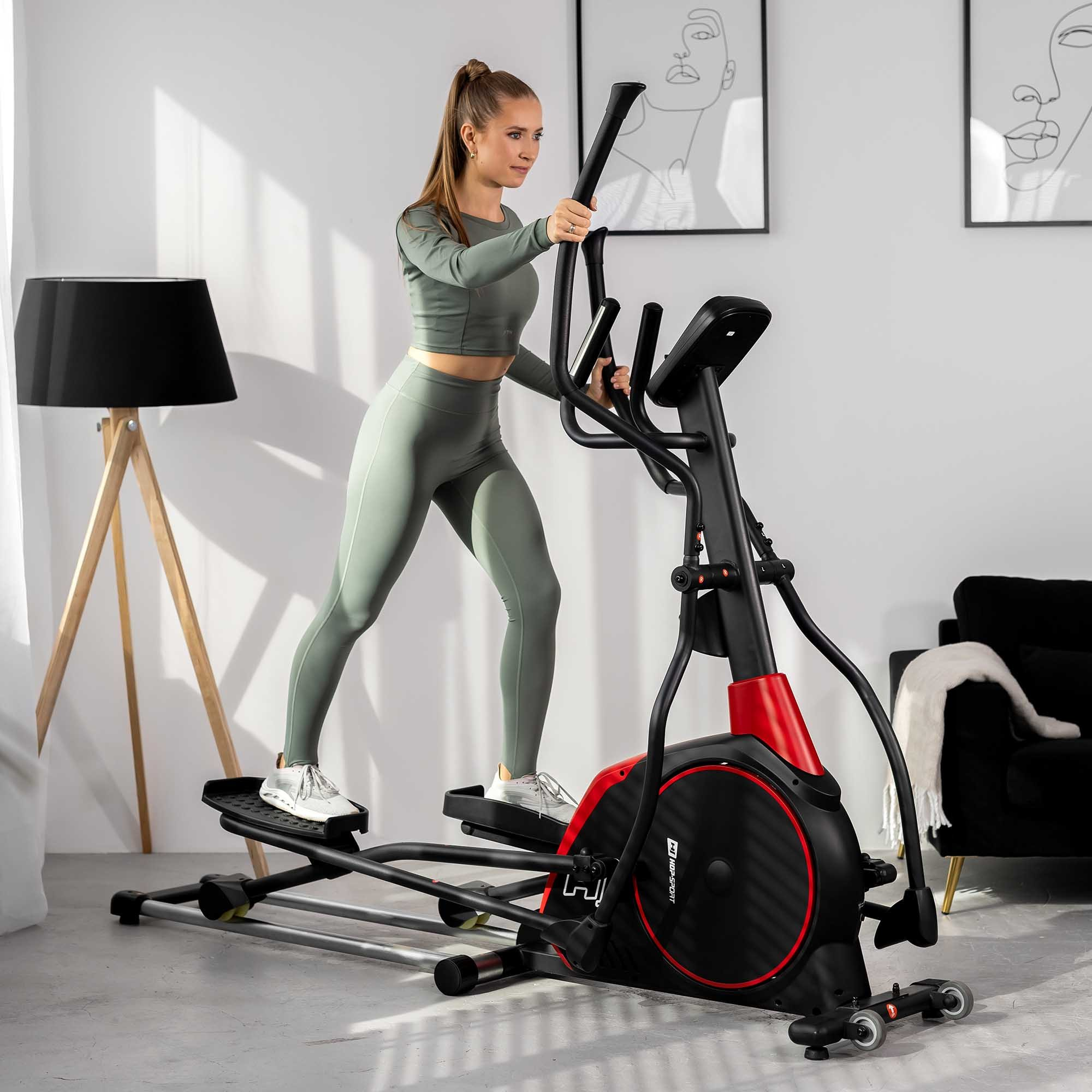 Eine Frau in Sportkleidung trainiert zu Hause auf einem schwarzen und roten Crosstrainer in einem hellen Raum.
