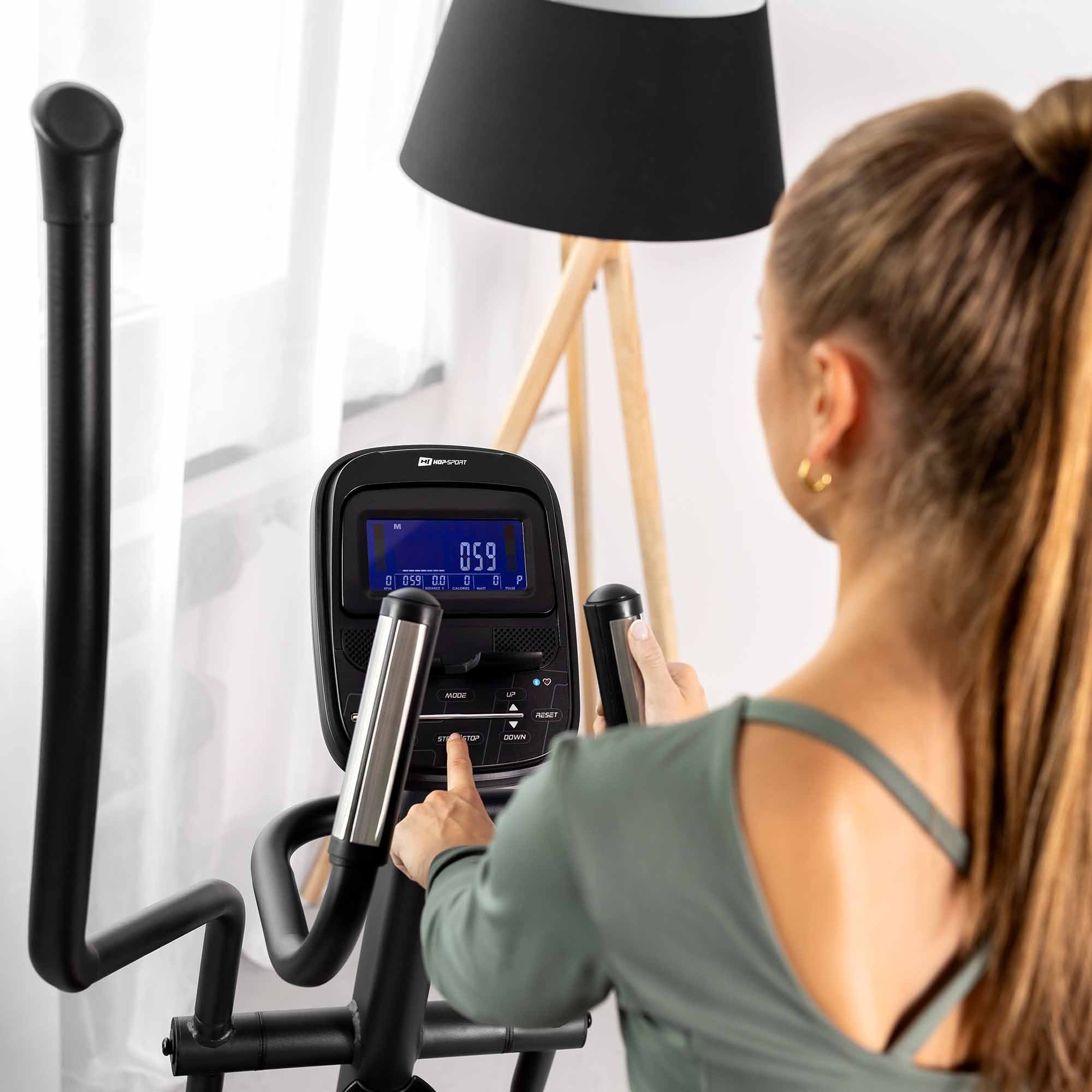Eine Frau im grünen Oberteil trainiert auf einem Crosstrainer und drückt in einem hellen Raum die Tasten.