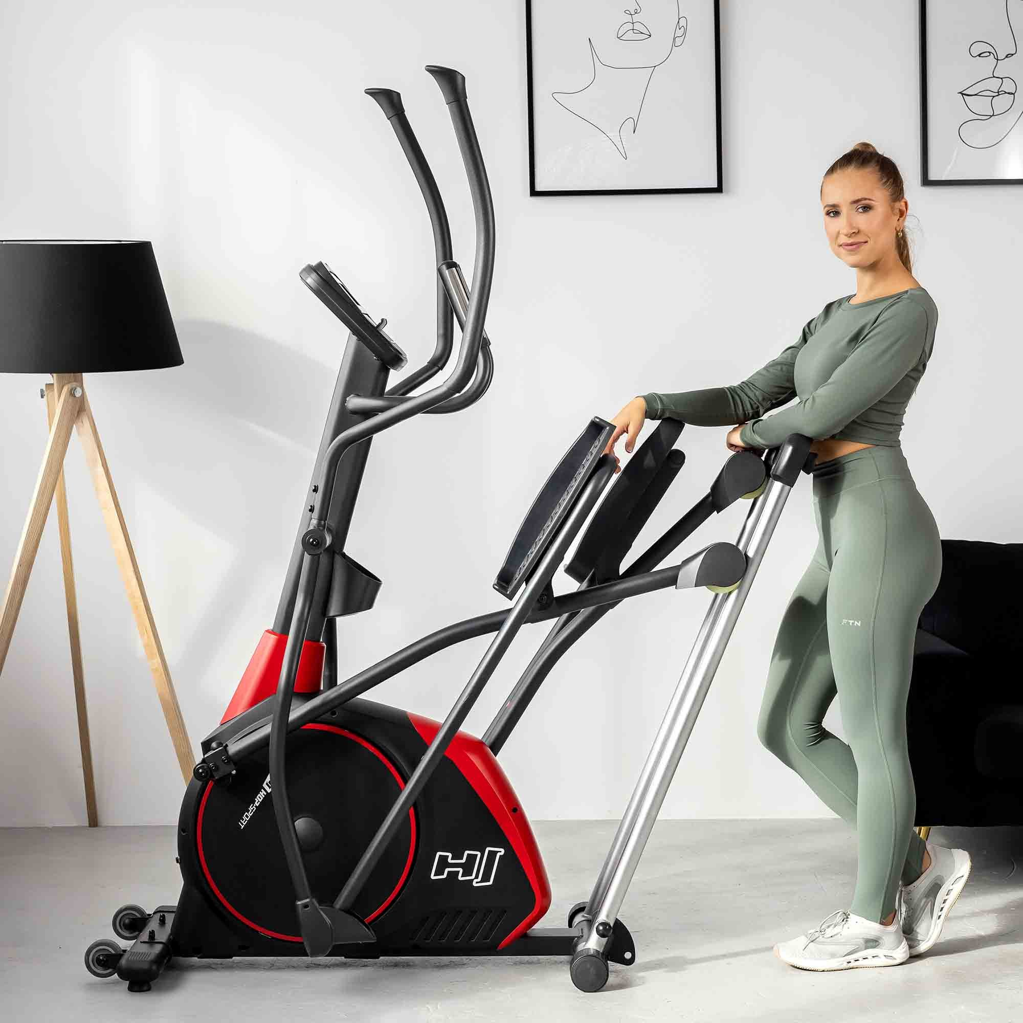 Eine Frau in grüner Sportkleidung posiert neben einem roten und schwarzen Crosstrainer in einem hellen Raum.