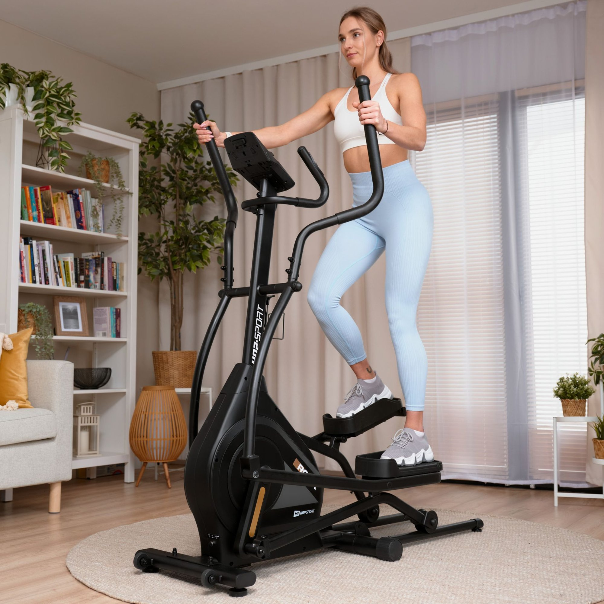 Frau im weißen Sport-Top und blauen Leggings trainiert zu Hause auf einem schwarzen Crosstrainer.