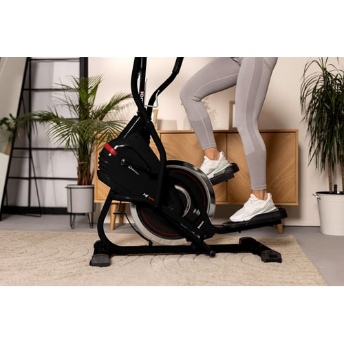 Eine Person trainiert zu Hause auf einem schwarzen Crosstrainer, umgeben von Zimmerpflanzen.