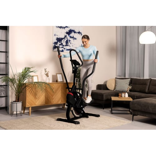 Eine Frau in Sportkleidung trainiert im Wohnzimmer auf einem schwarzen Crosstrainer.