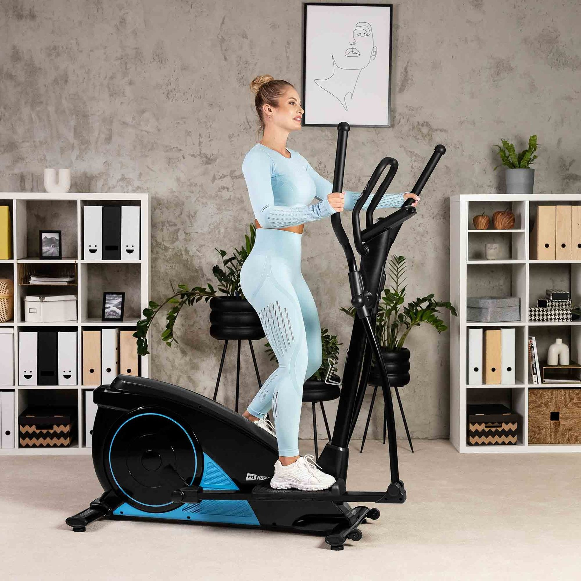 Eine Frau in blauer Sportkleidung trainiert zu Hause auf einem schwarzen Crosstrainer vor einem Regal.