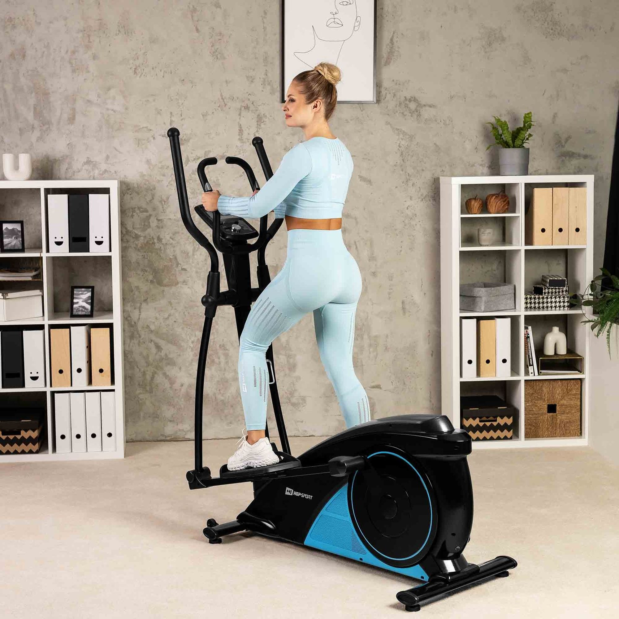 Frau in hellblauer Sportbekleidung trainiert auf einem schwarzen und blauen Crosstrainer in einem modernen Zimmer.