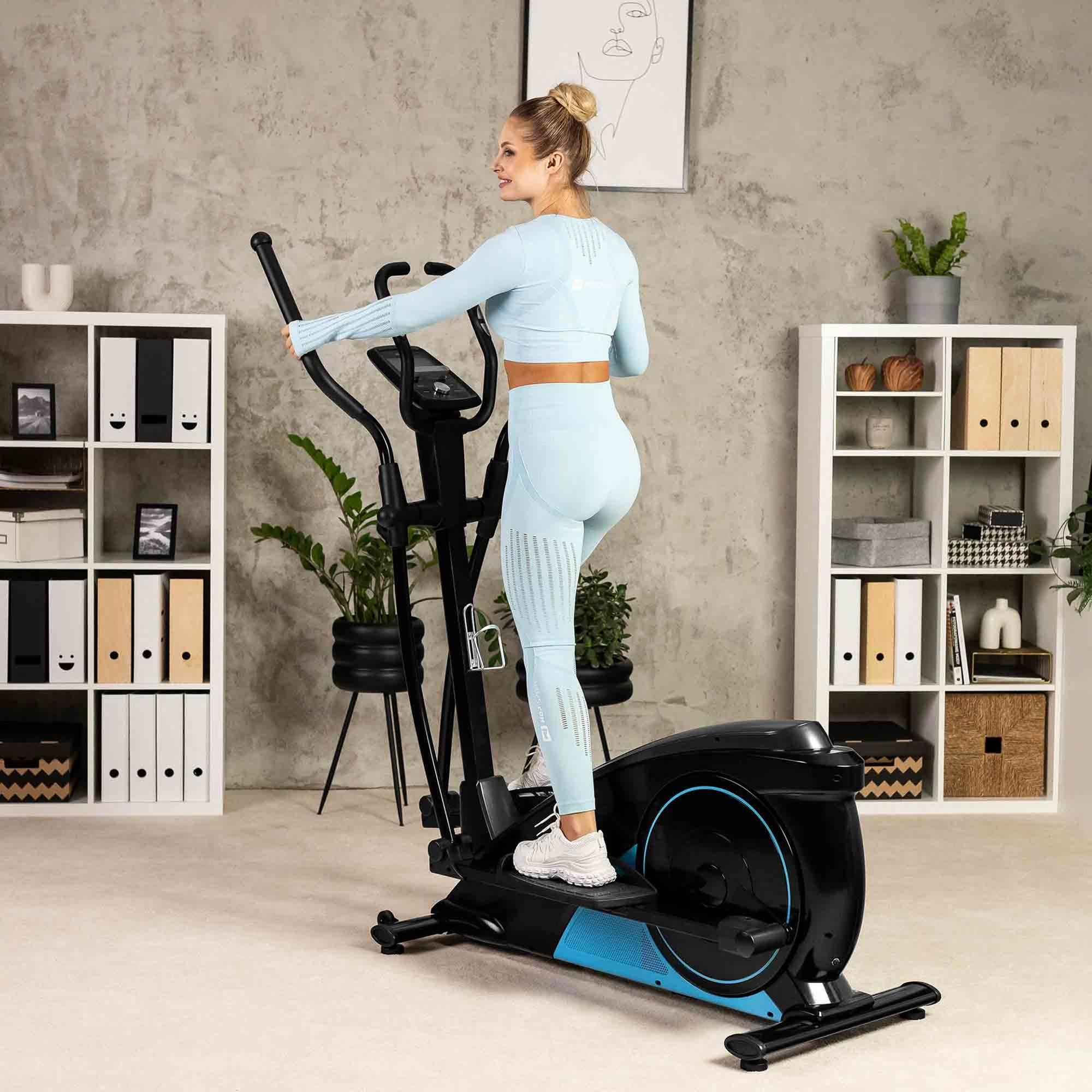 Eine blonde Frau in blauer Sportkleidung trainiert auf einem schwarz-blauen Crosstrainer in einem hellen Raum.